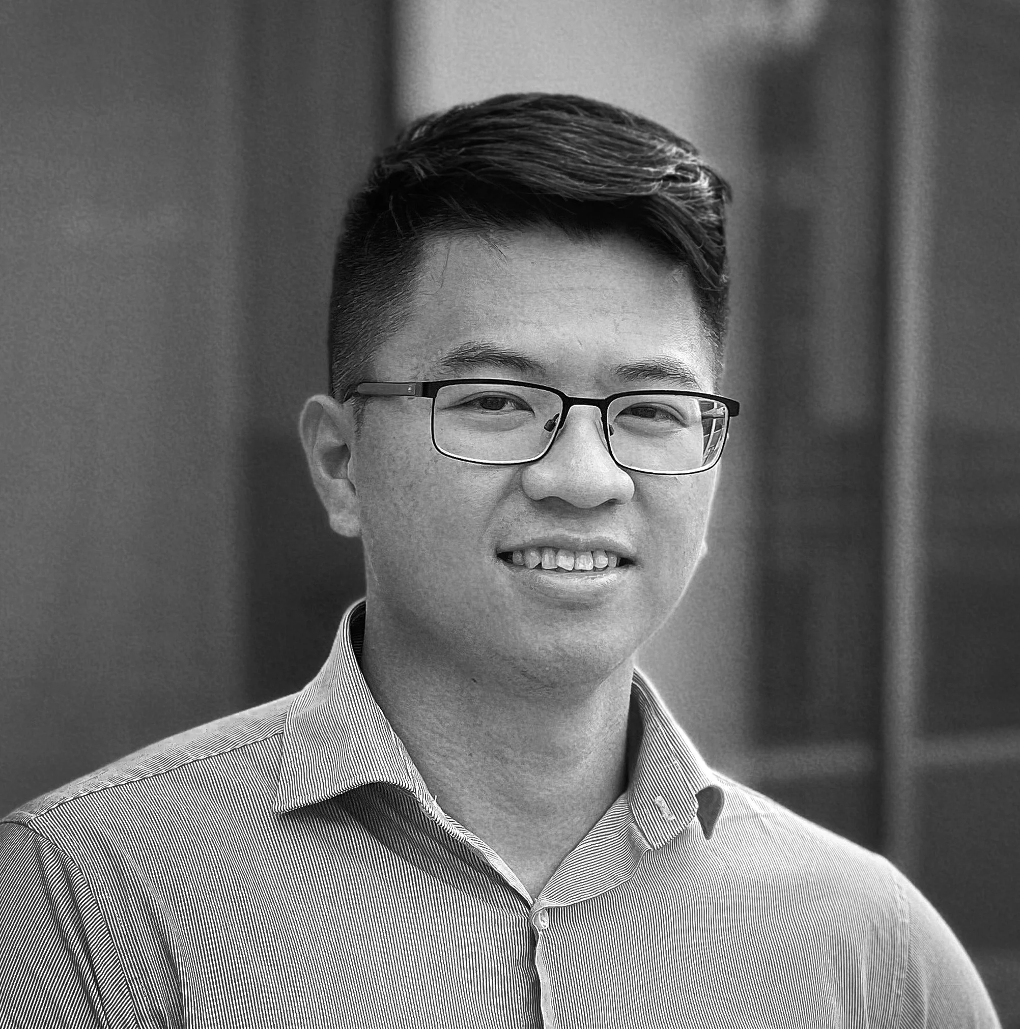 A young man with short dark hair and glasses, smiling, wearing a light blue collared shirt, standing outdoors with a modern glass building in the background.