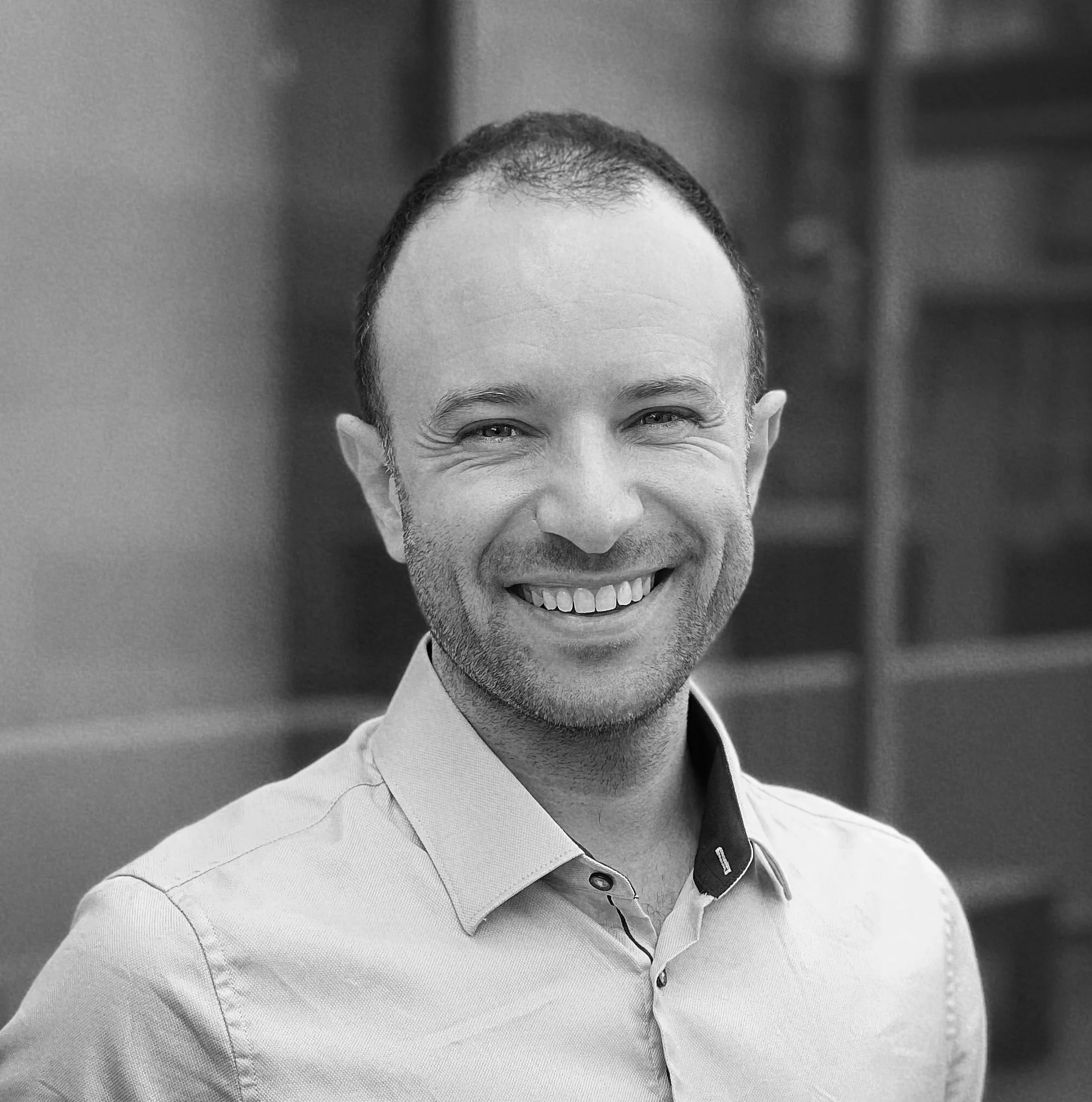 Smiling man with short dark hair and a beard, wearing a light blue collared shirt, standing outside near glass building reflects, in an urban setting.