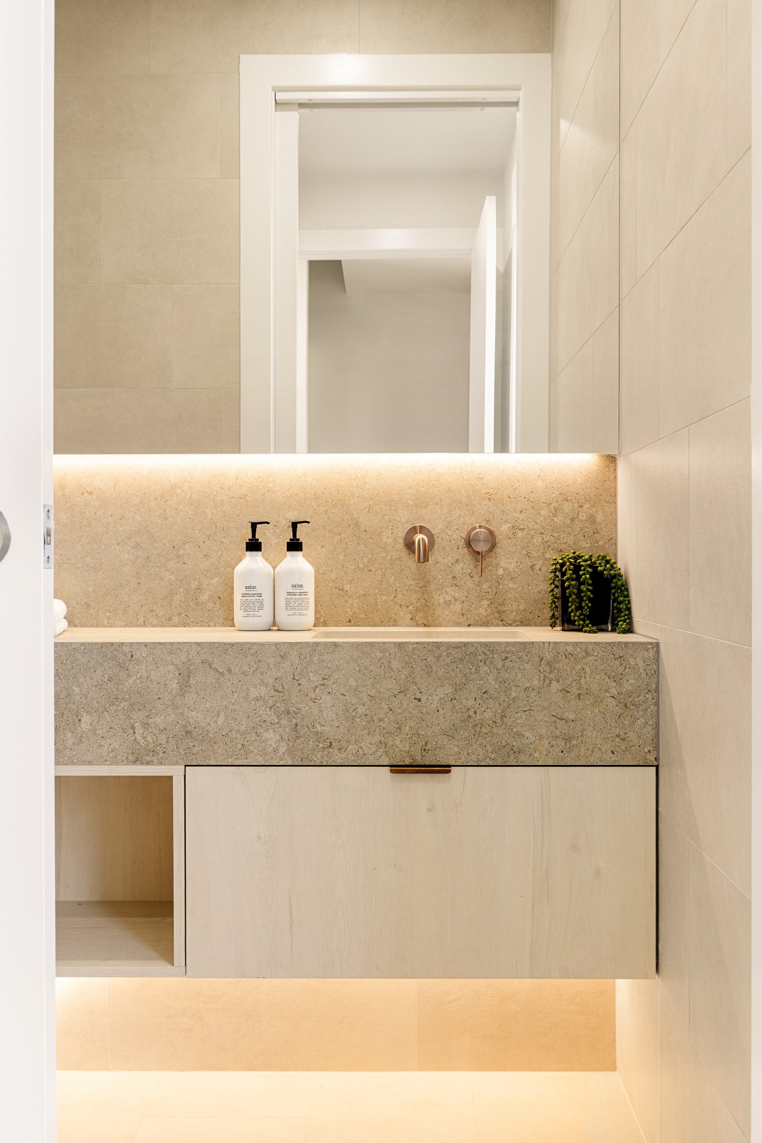 A bathroom vanity with a beige countertop holding two soap dispensers and a small green potted plant. The mirror above reflects a hallway with white walls and a doorframe.
