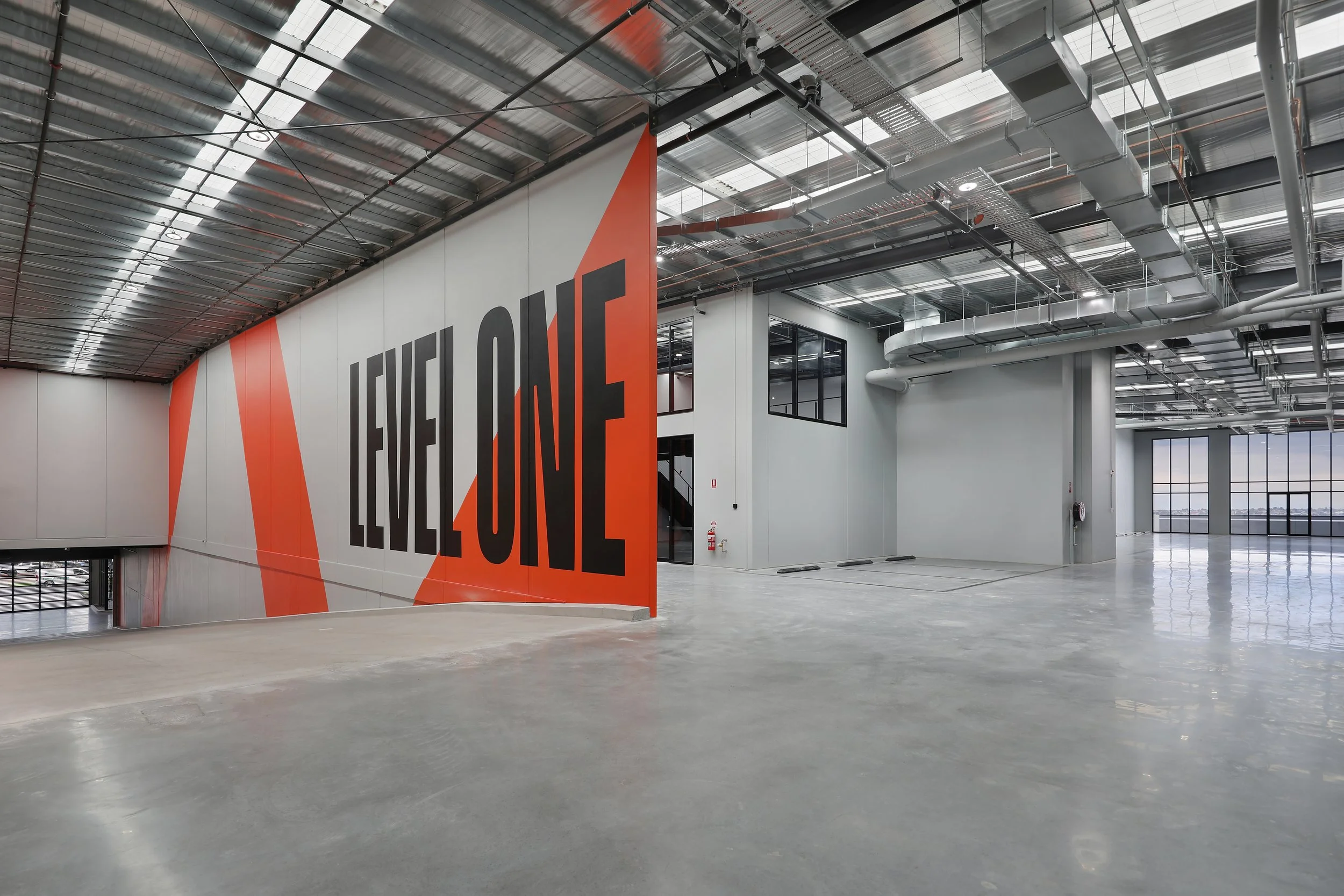 Modern interior of a large industrial building or warehouse with a high metal ceiling, exposed pipes and ducts, large windows, and a big wall with a sign that reads 'FUELLINE' in bold black letters on a red and white background.