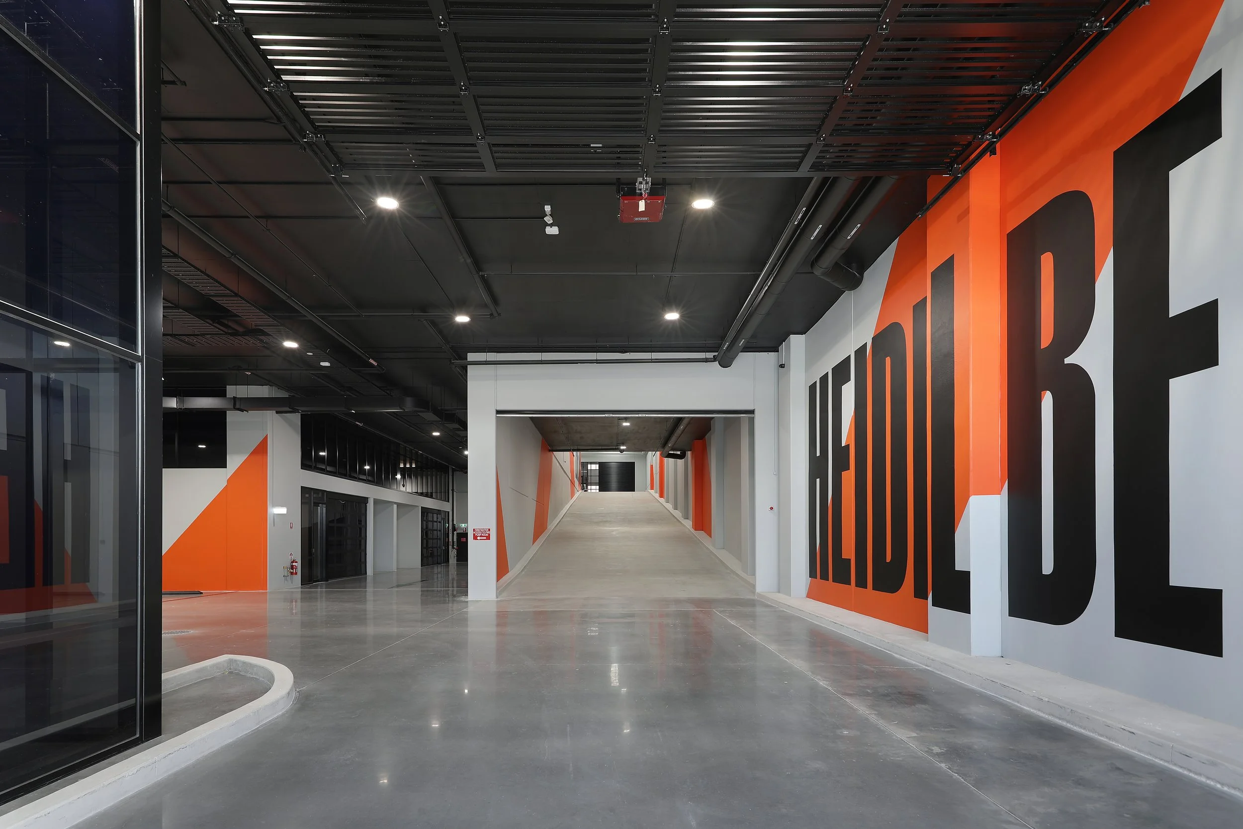 Brightly lit indoor corridor with black and orange wall art, sleek concrete floor, and visible ceiling piping.