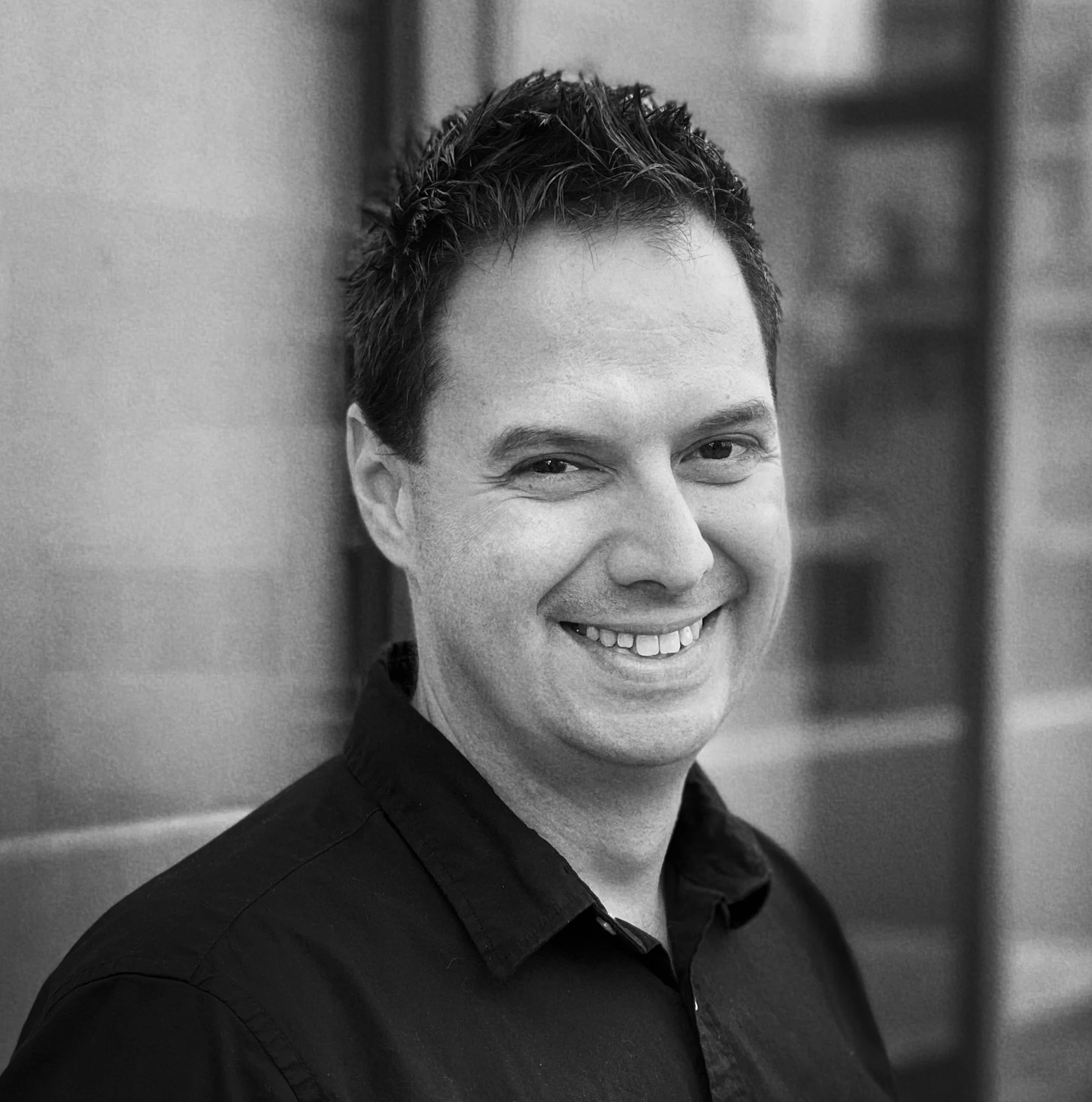 Smiling man with short dark hair wearing a black shirt, standing outdoors with a blurred blue building in the background.