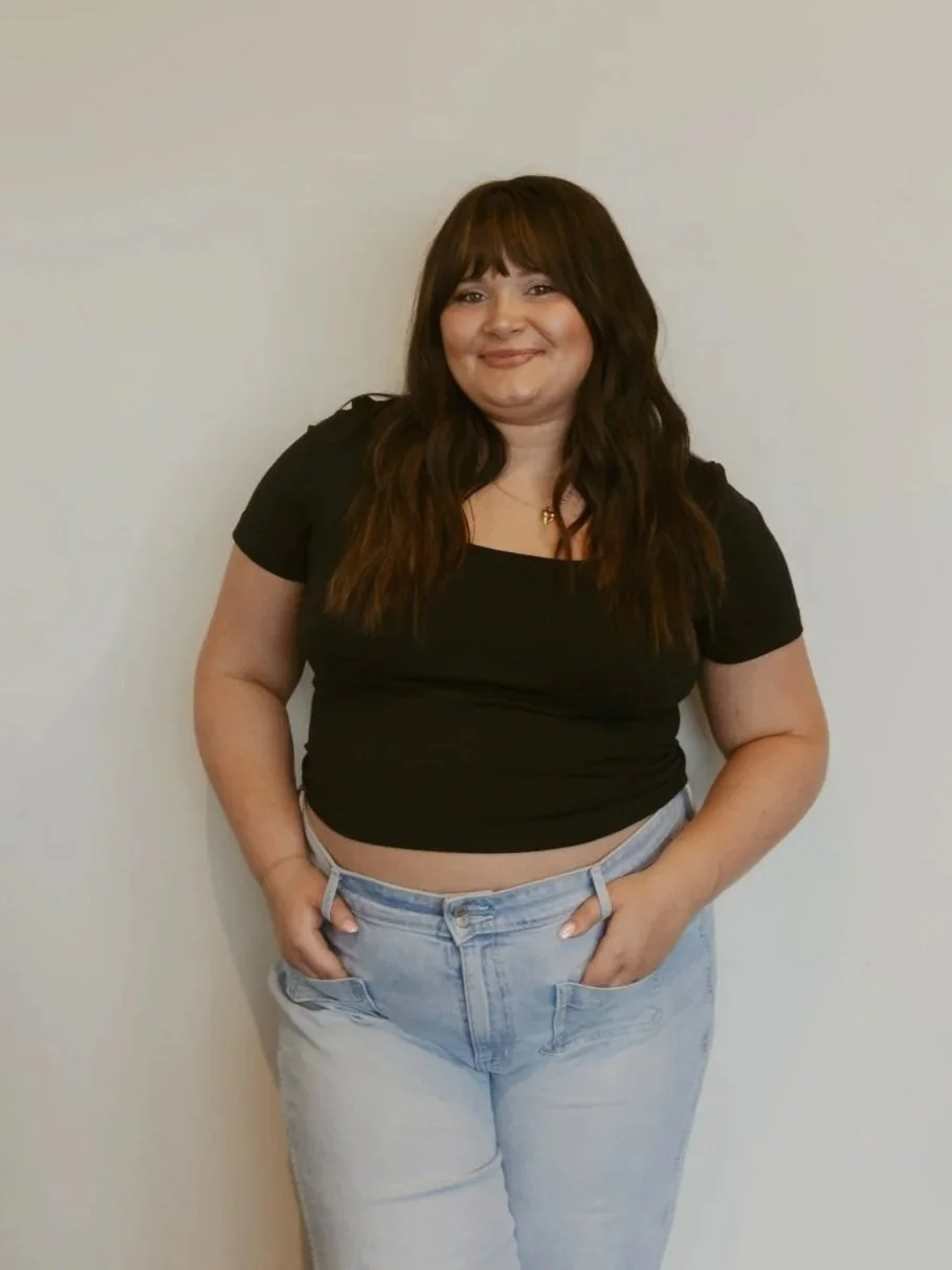 A woman with long brown hair and bangs, smiling and wearing a black crop top and light blue jeans, standing against a plain off-white wall.