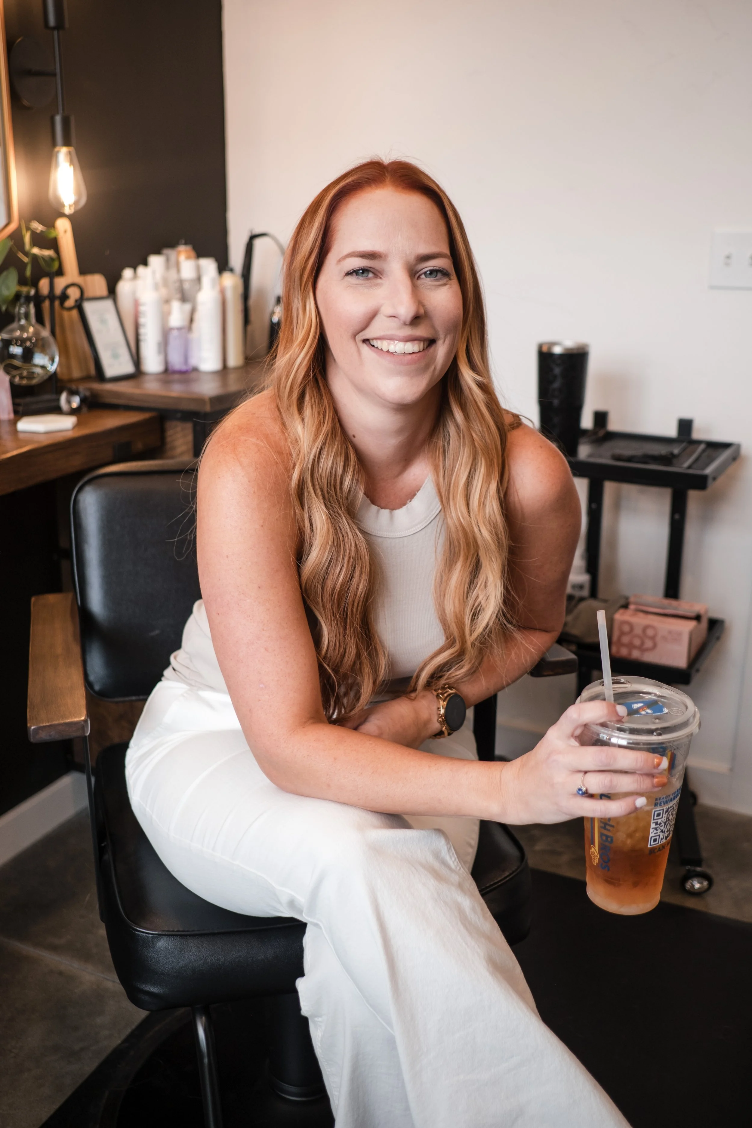 A woman with long wavy red hair smiling and sitting in a salon or spa, holding a large iced beverage in a clear plastic cup with a straw, wearing a white sleeveless top and white pants.