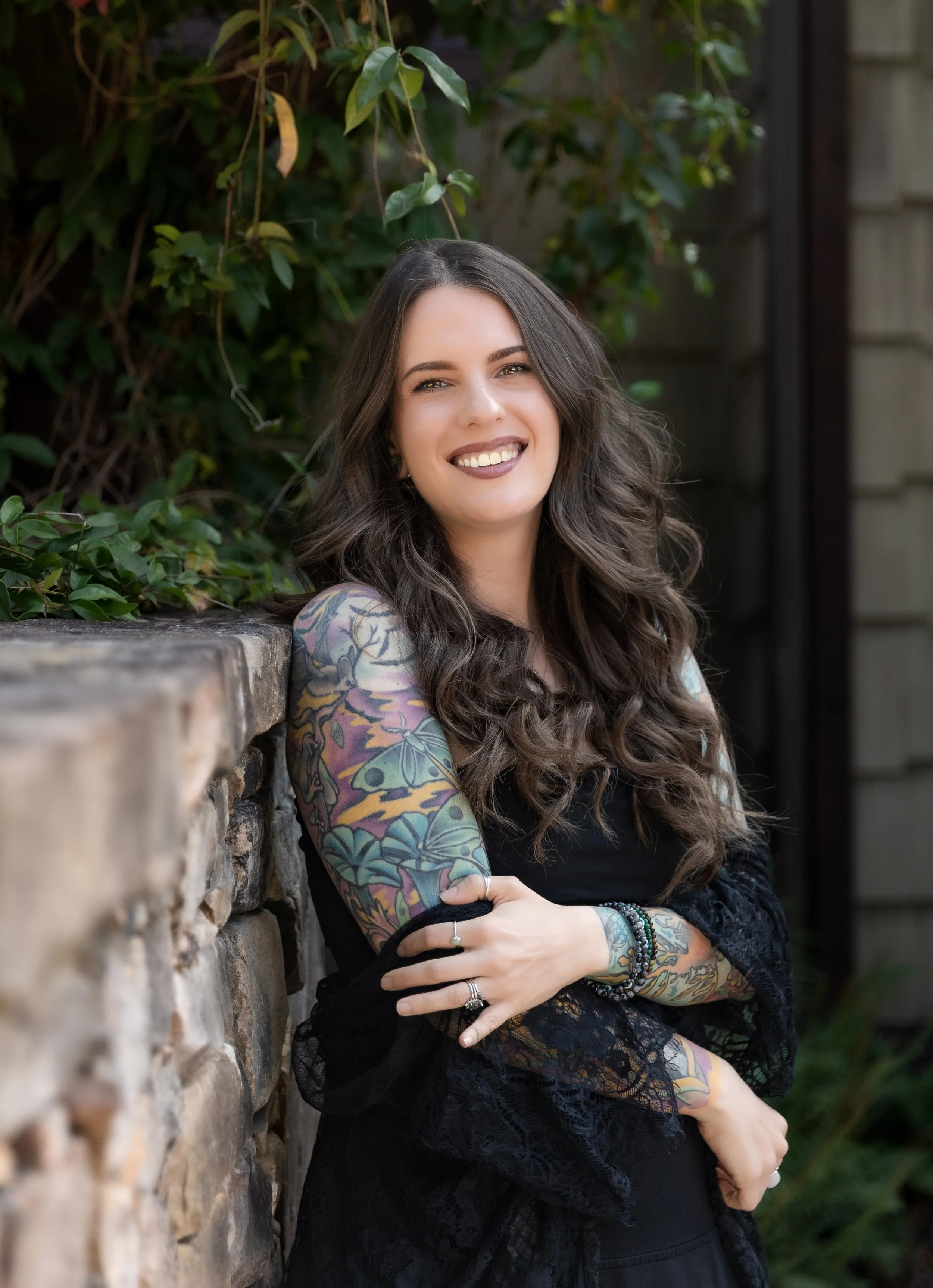 Courtney, a woman with long brown hair and tattoos in a lace shawl and all black clothing, which aligns with her saturn placement smiles for the camera.