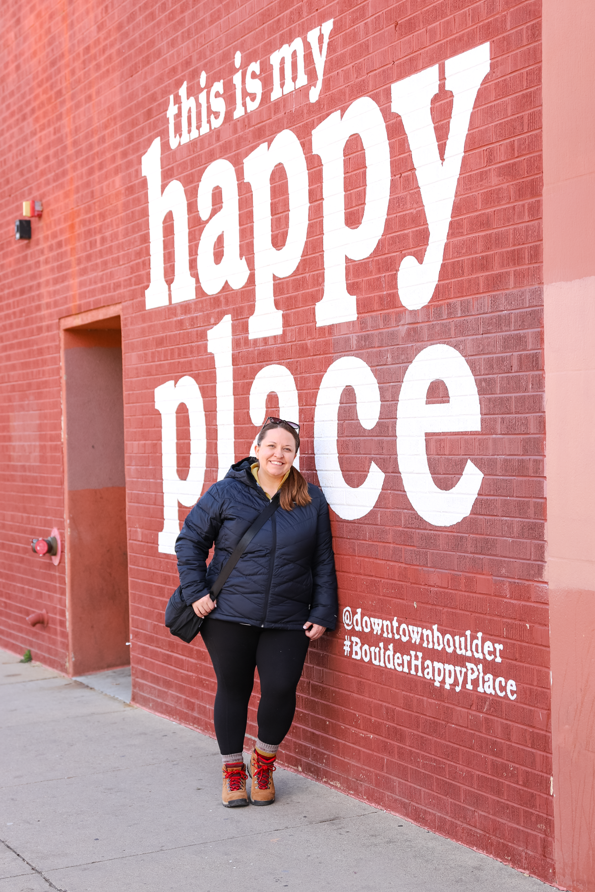 Woman smiling and leaning against a brick wall with the text 'this is my happy place' and hashtags @downtownboulder, #BoulderHappyPlace.