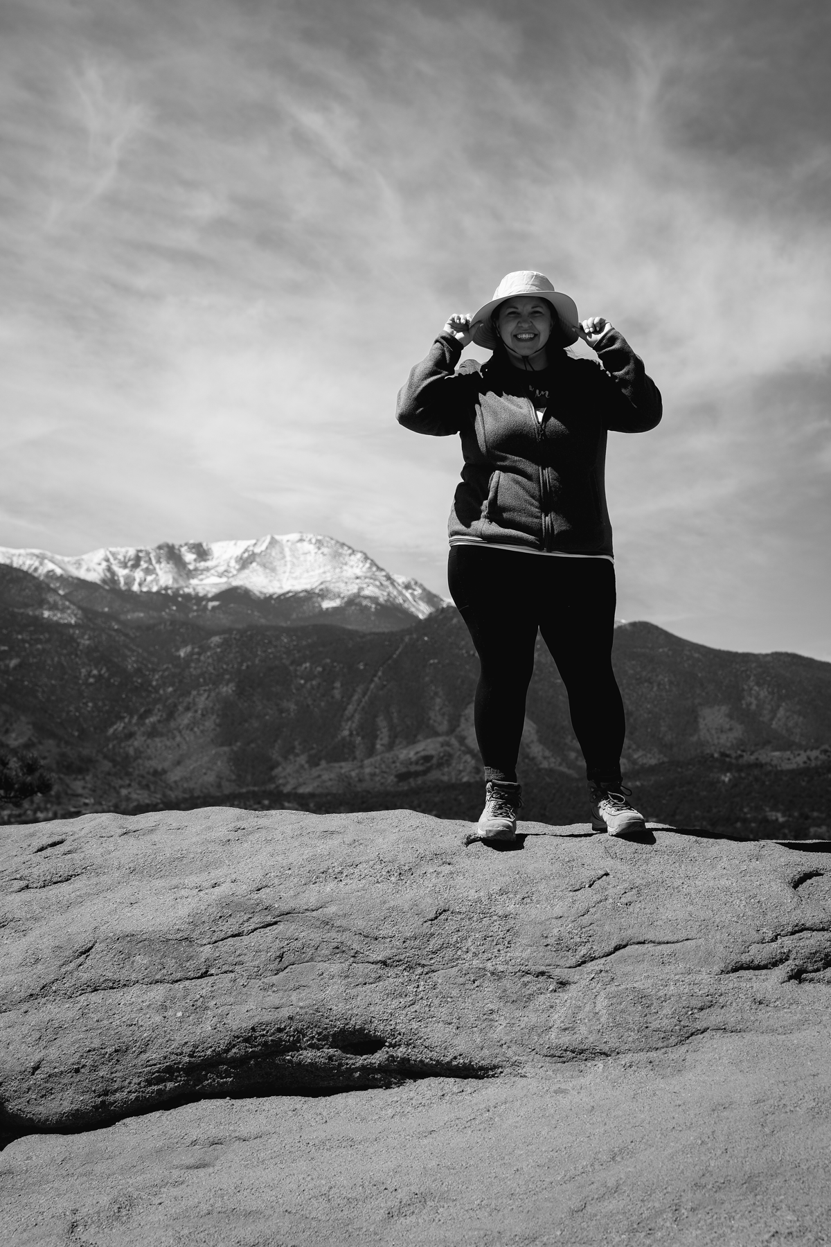 Person standing on a rock with mountains in the background, wearing a hat and outdoor clothing.