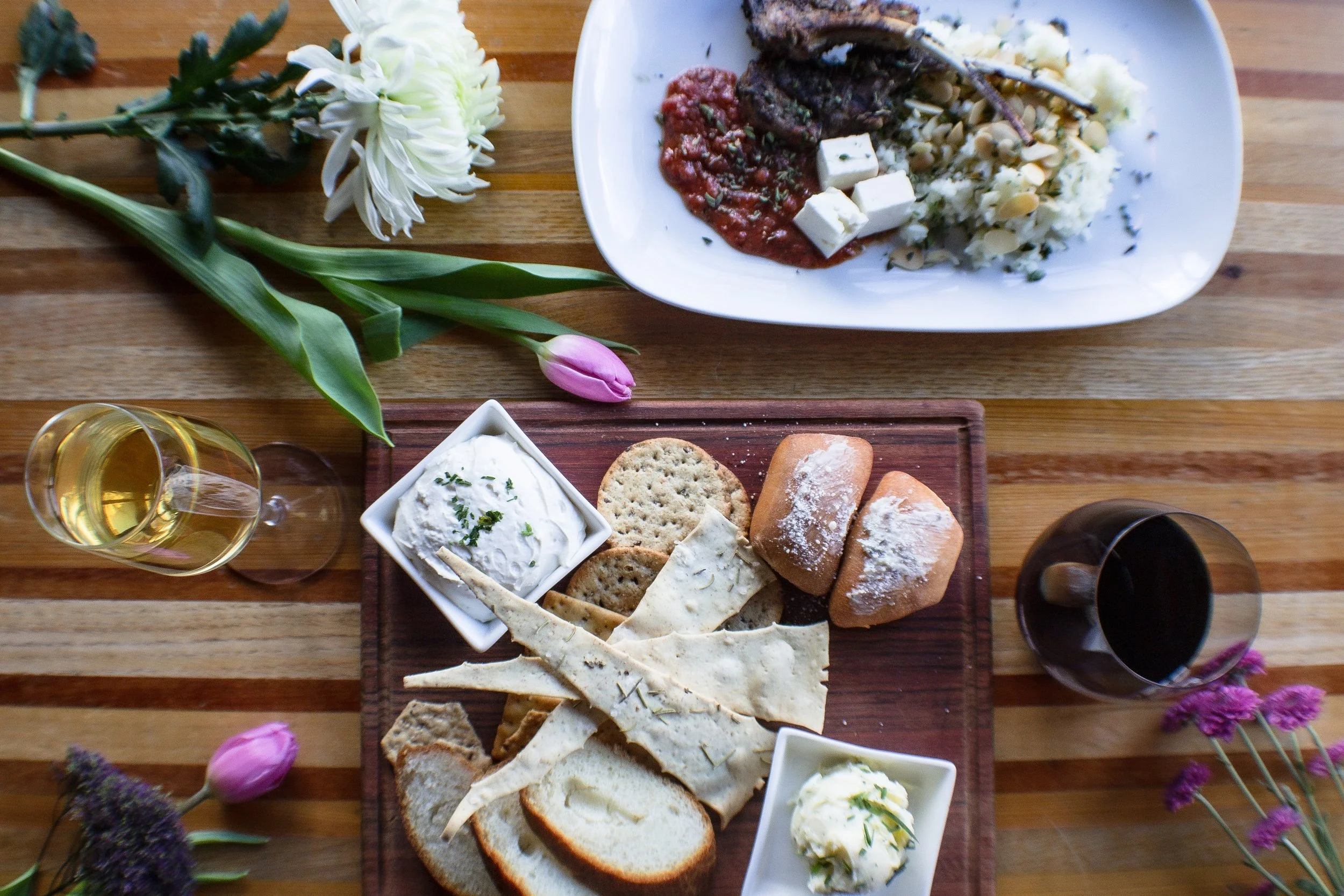 A dining table setting with a plate of grilled lamb chops, mashed potatoes, feta cheese, and tomato sauce. There is a wooden board with a selection of bread, crackers, and cheese spread. There are glasses of white and red wine, and floral decorations including white and purple flowers.