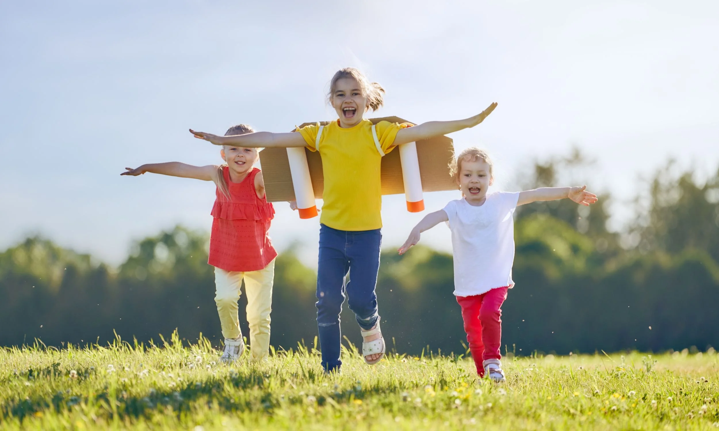 Three happy young girls running in a sunlit field, pretending to fly with cardboard wings.