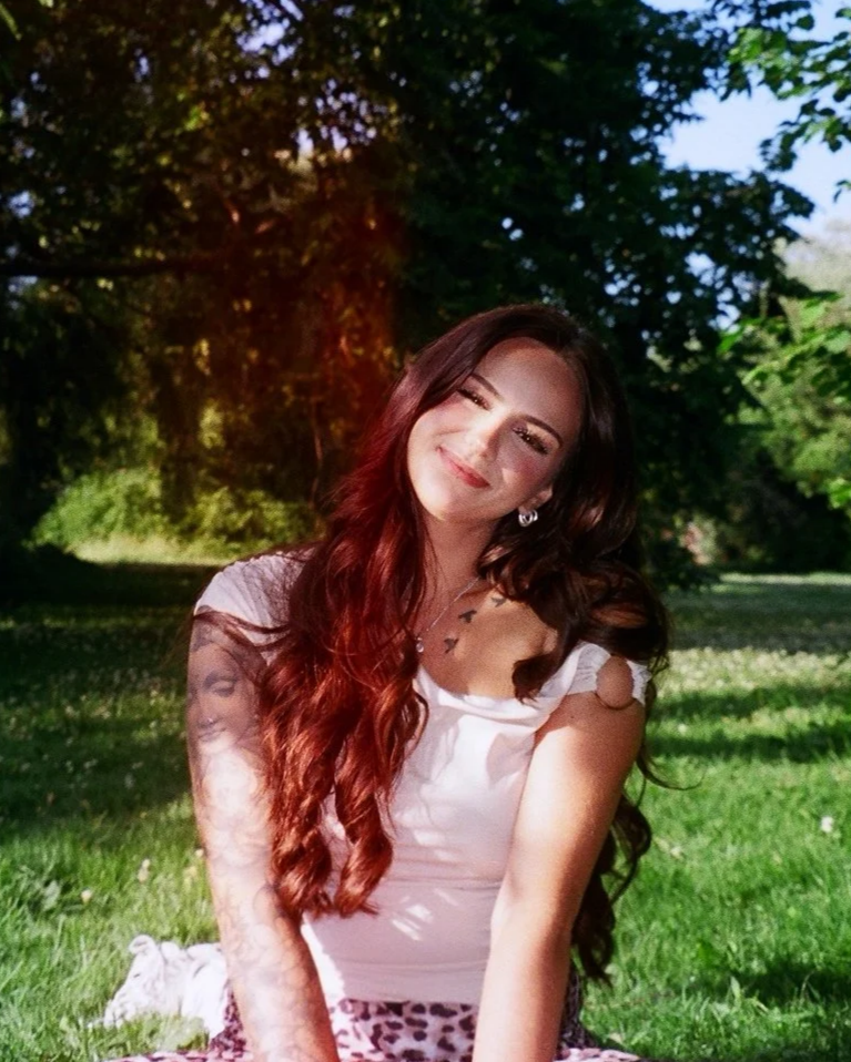 A young woman with long, wavy red hair sitting outdoors on grass, smiling with her eyes closed, wearing a white top and leopard print pants, with trees and sunlight in the background.