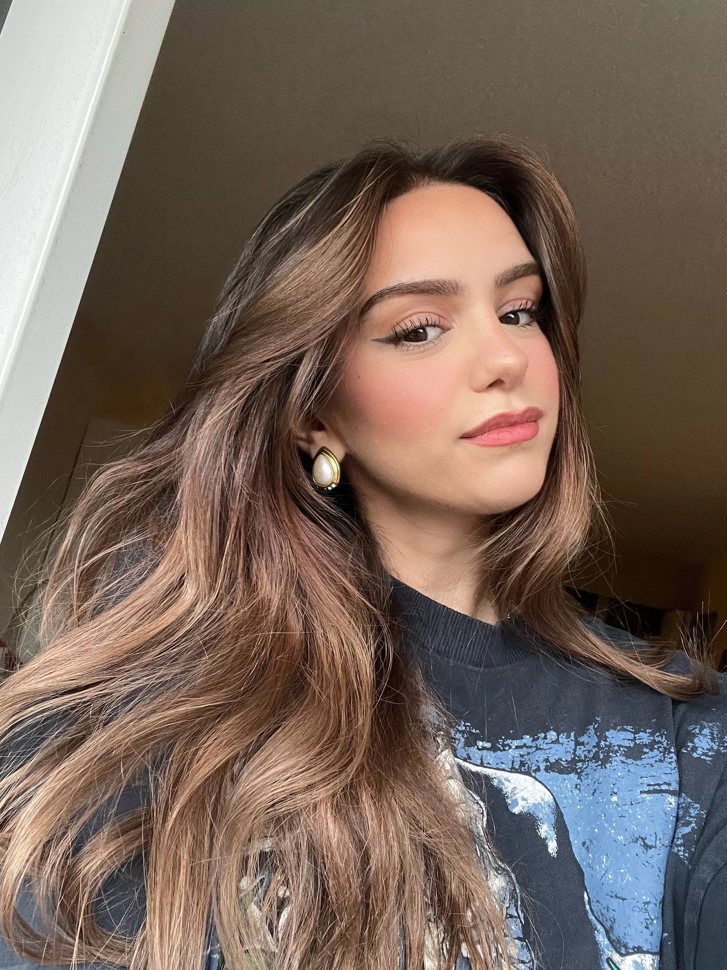 A young woman with long, wavy brown hair and earrings, wearing a black graphic shirt, is posing for a selfie indoors.