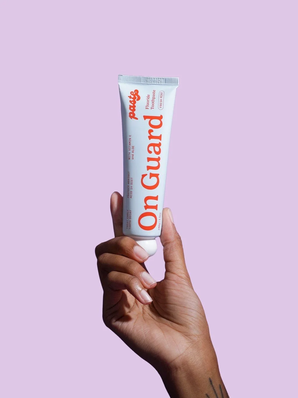 A hand holding a tube of mint fluoride toothpaste with a violet background..