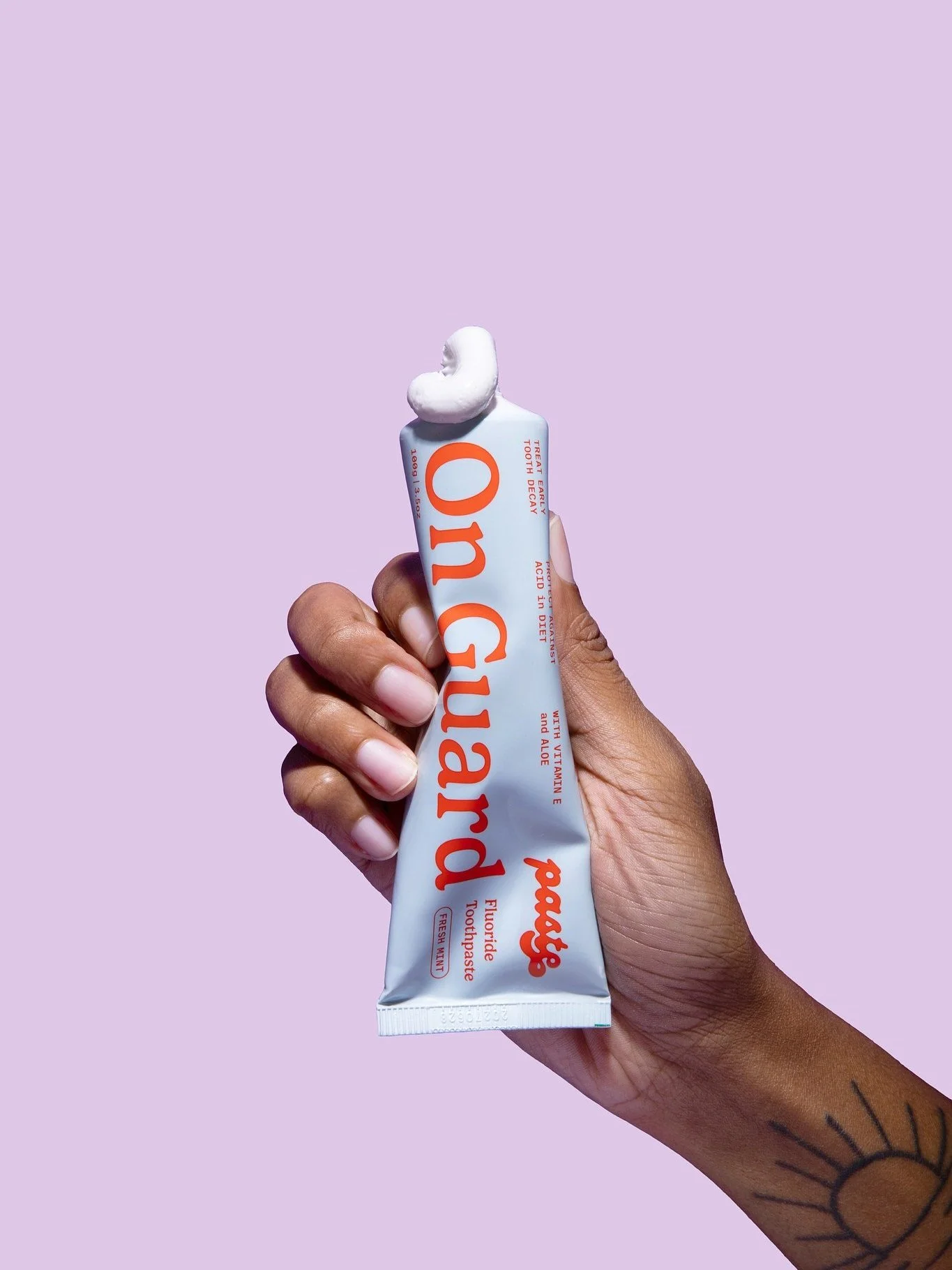 A hand squeezing a tube of mint fluoride toothpaste with a violet background..