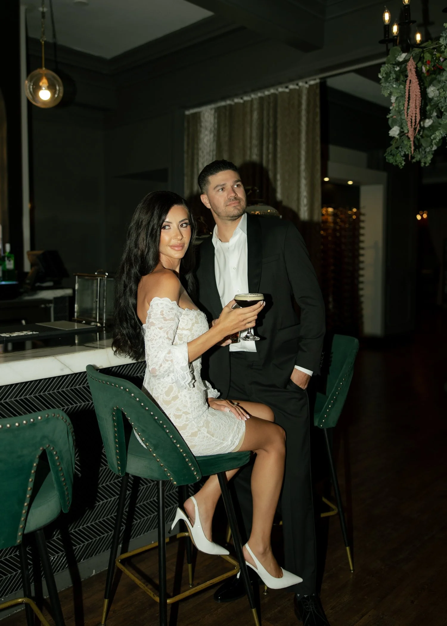 couple in white dress and black suit with a nostalgic film looking golding a cocktail not looking at the camera