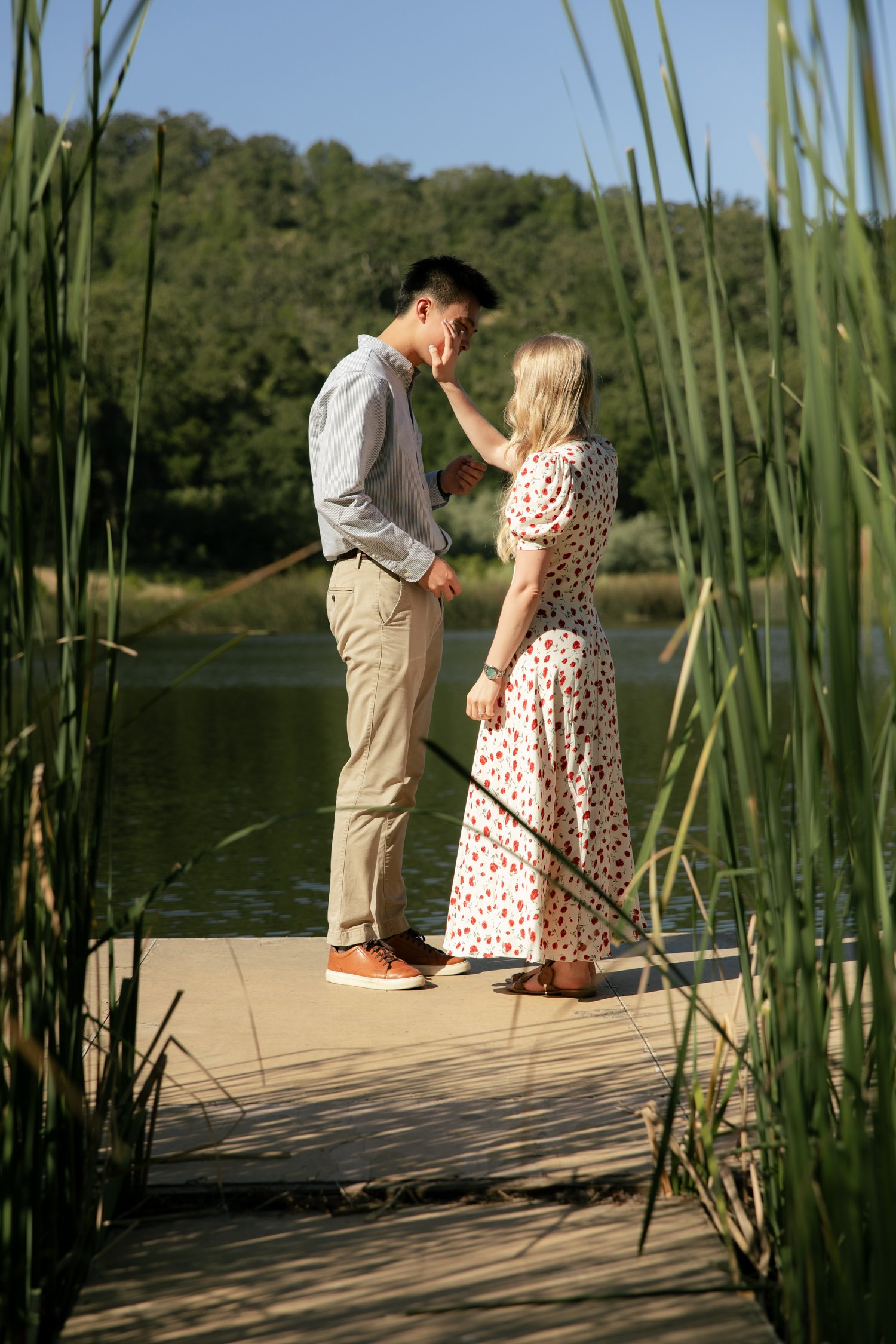two people standing on a dock as one wipes away tears.they are surrounded by reeds with a lake behind them