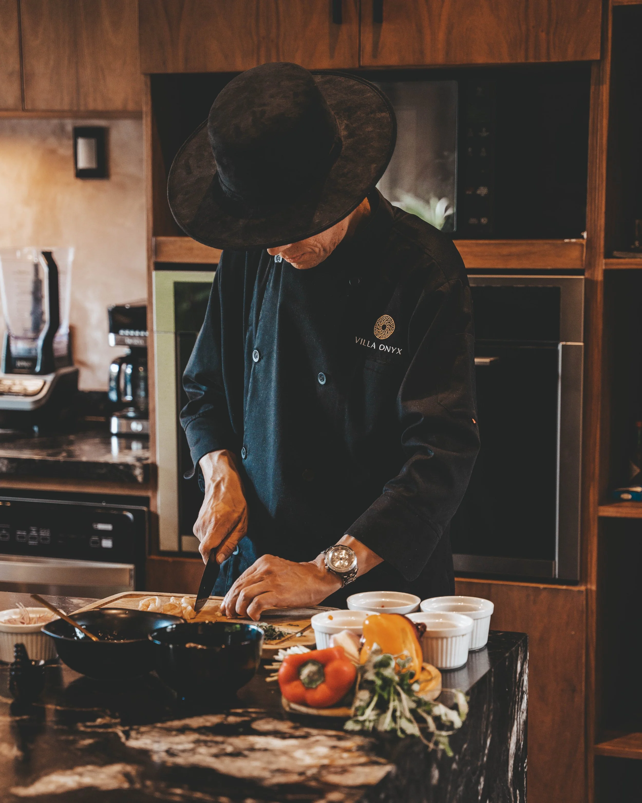 Man in black chef's uniform with Villa Onyx logo slicing food in a kitchen with wooden cabinets, surrounded by various dishes and ingredients.