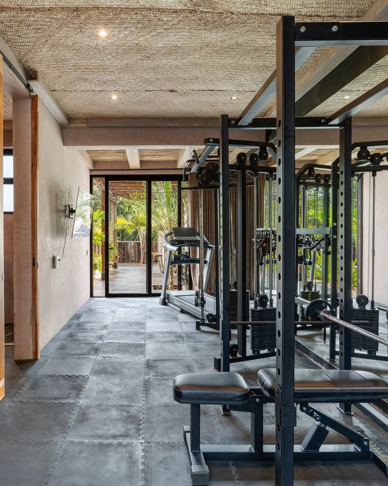 Indoor gym with fitness equipment including a treadmill, weight rack, and benches, with large glass doors leading to an outdoor patio with greenery.