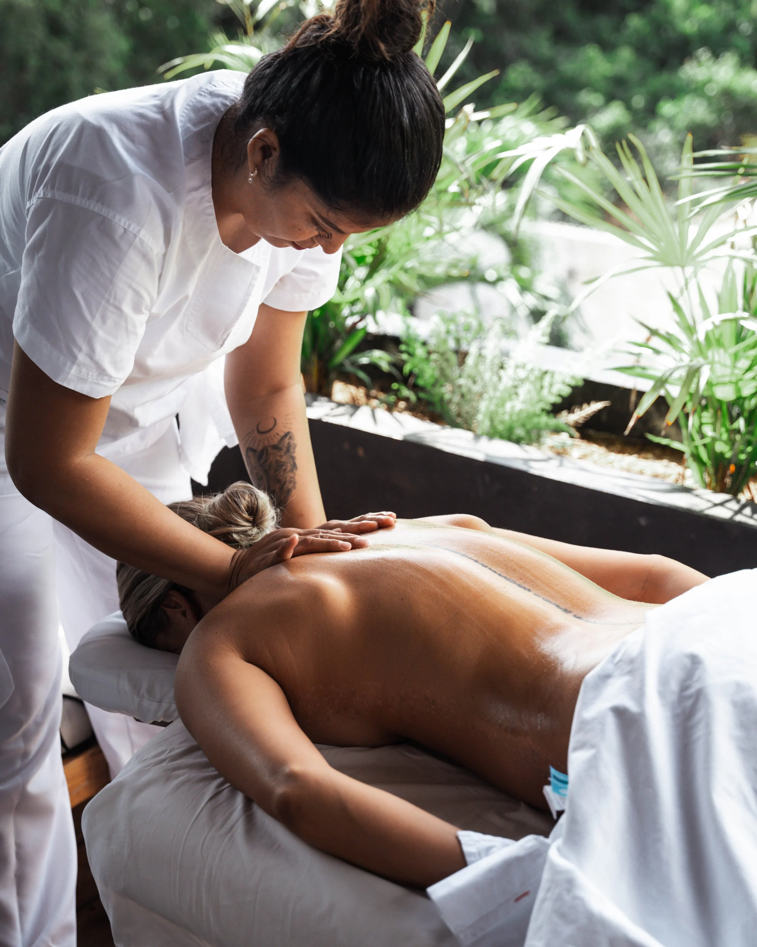 A person receiving a massage outdoors, lying face down on a massage table with a white sheet, surrounded by green plants.