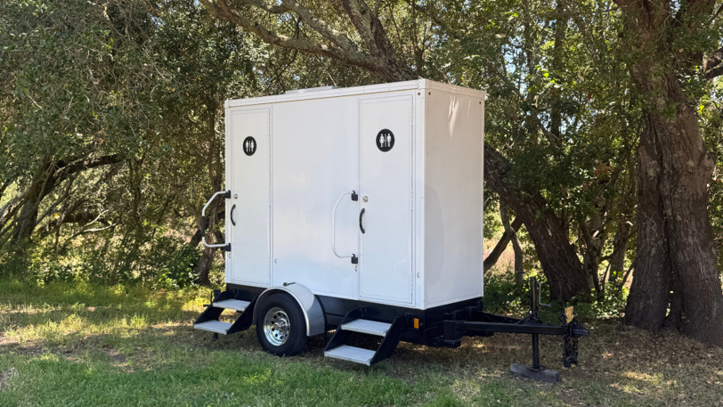 A luxury restroom trailer with two stalls set up on a grassy field near trees.