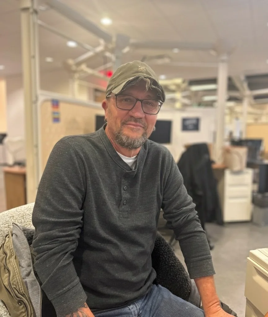 A man wearing glasses, a baseball cap, and a gray short-sleeve button-up shirt standing outside in front of a tree and brick building.