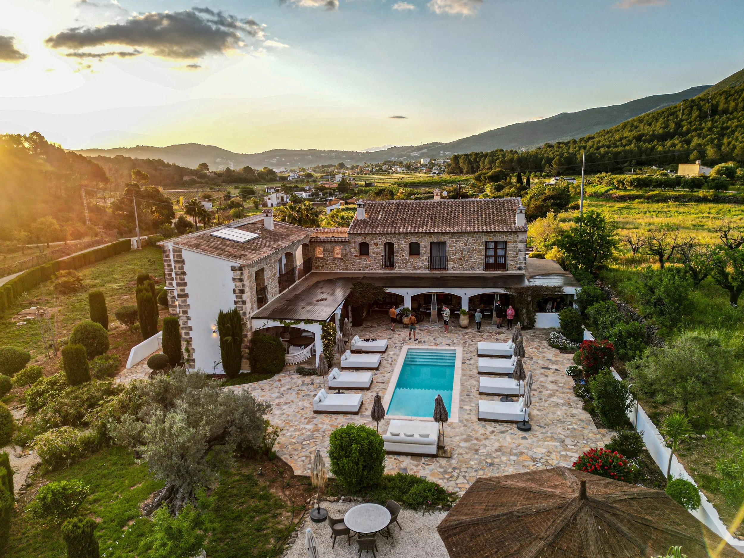 A rustic two-story house with a stone facade and tiled roof, surrounded by a landscaped garden with a swimming pool and lounge chairs, set in a scenic countryside with rolling hills and fields in the background during sunset.