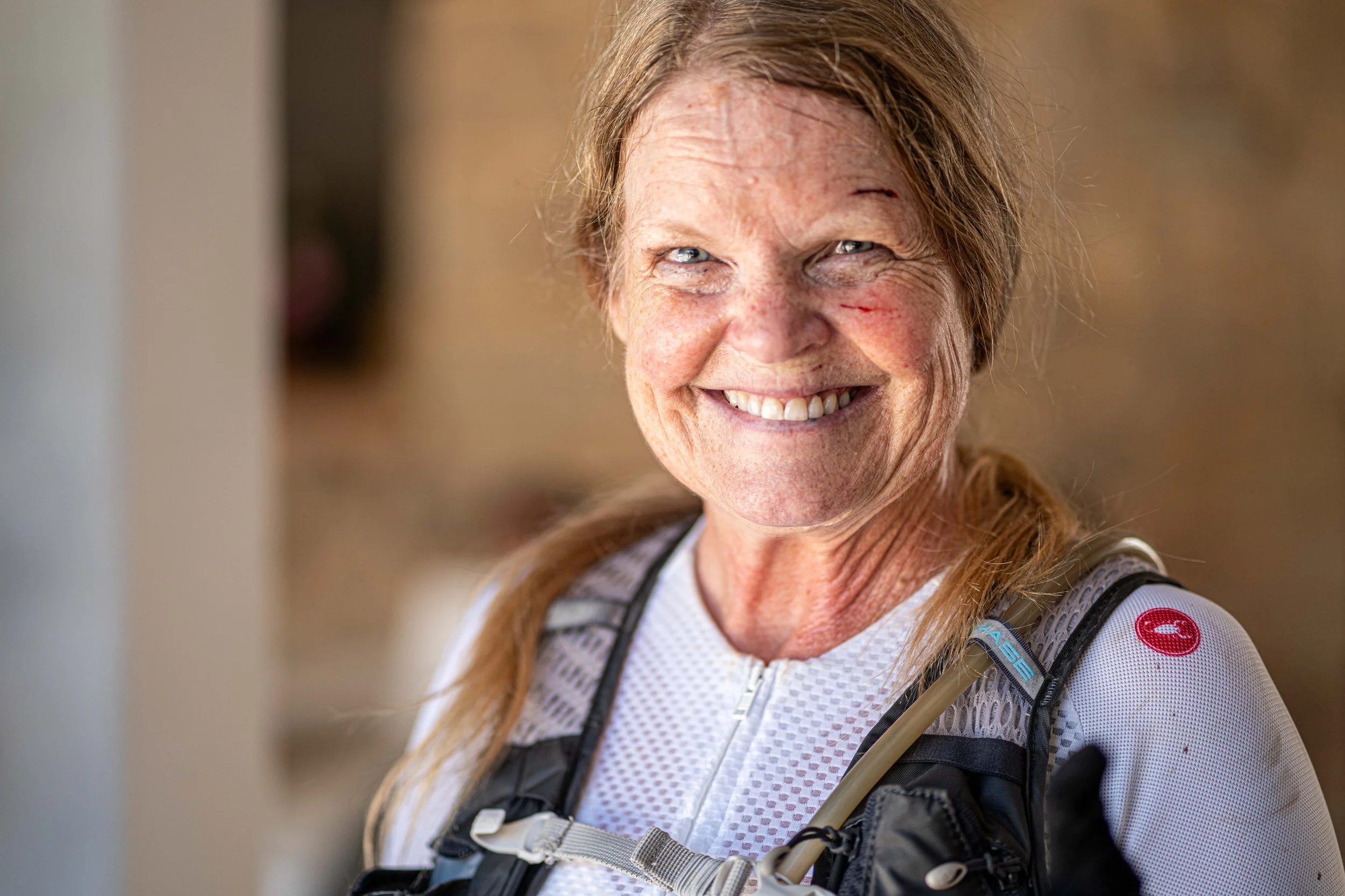 Close-up of a smiling senior woman with freckles, wearing a white athletic top and a hydration backpack.