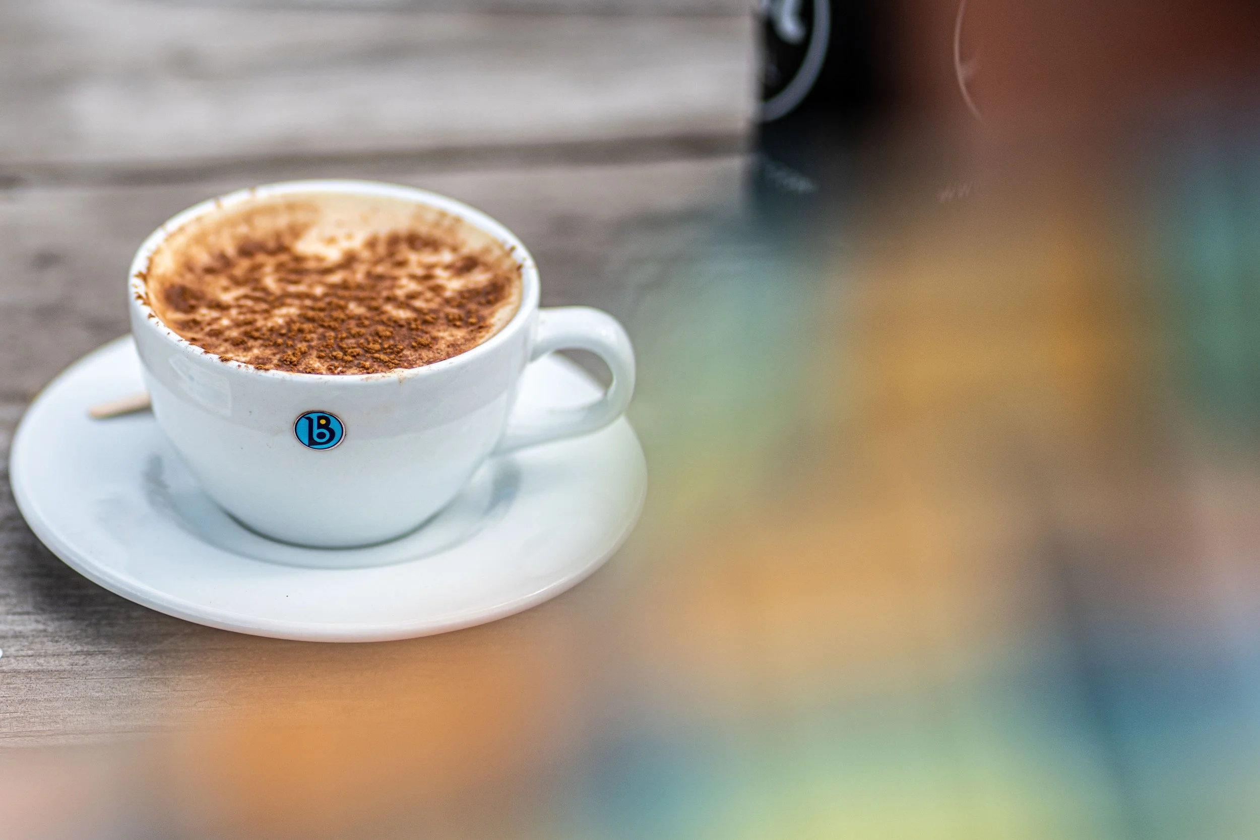 A white cup of coffee at Blanca Bikes, with foam and cocoa powder on top, sitting on a matching saucer on a wooden outdoor table.
