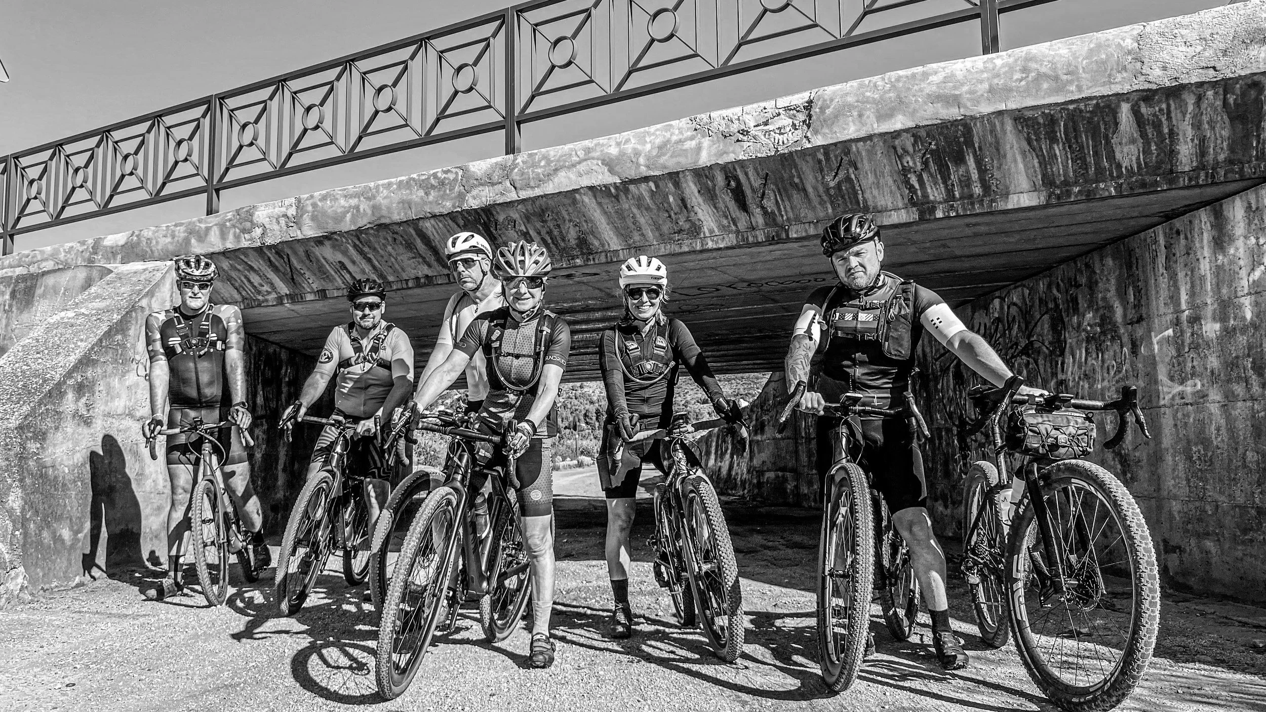 Six cyclists in cycling gear and helmets with mountain bikes under a bridge.