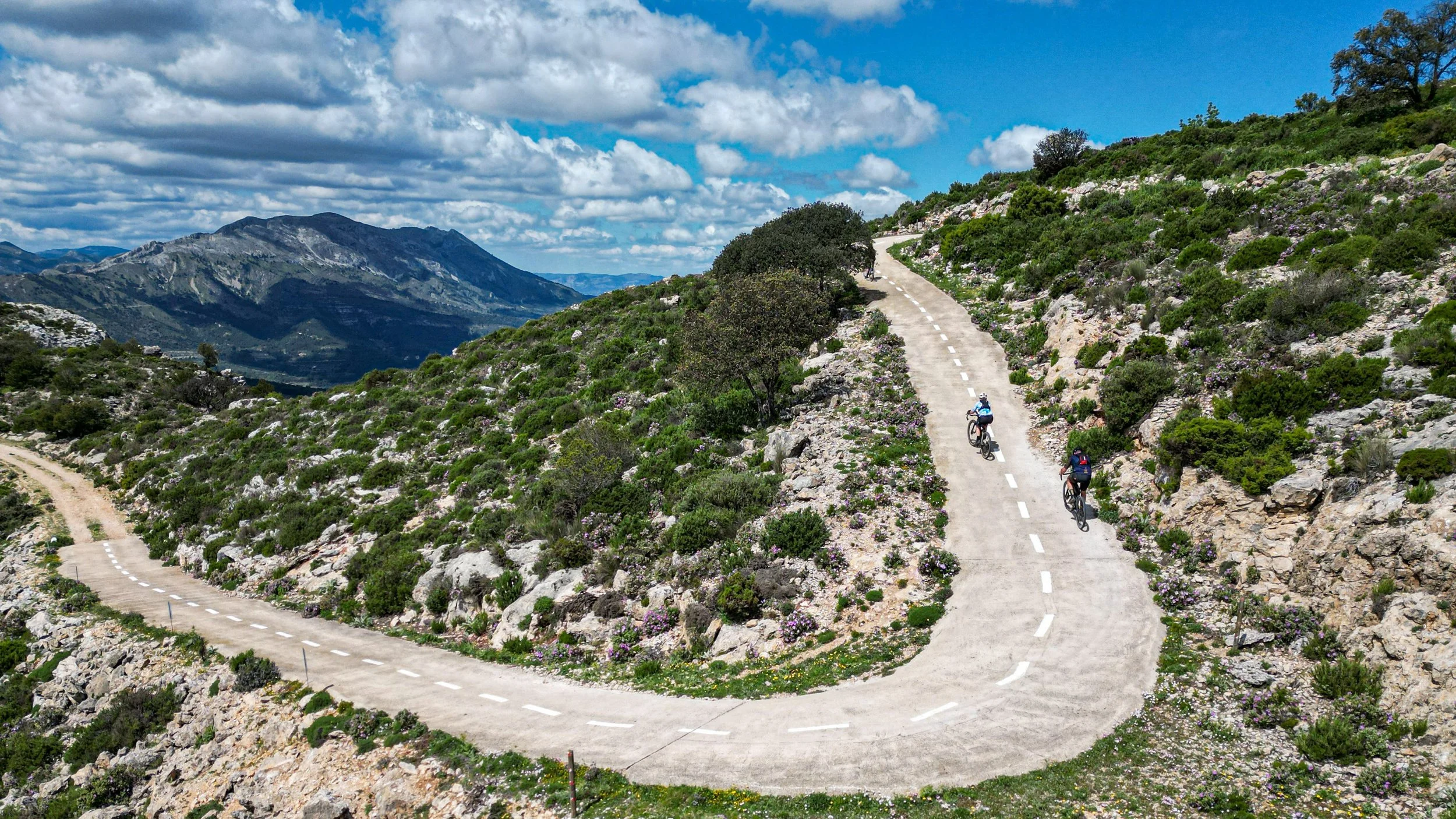 Two cyclists riding on a winding mountain trail with scenic mountain landscape and cloudy sky in the background.