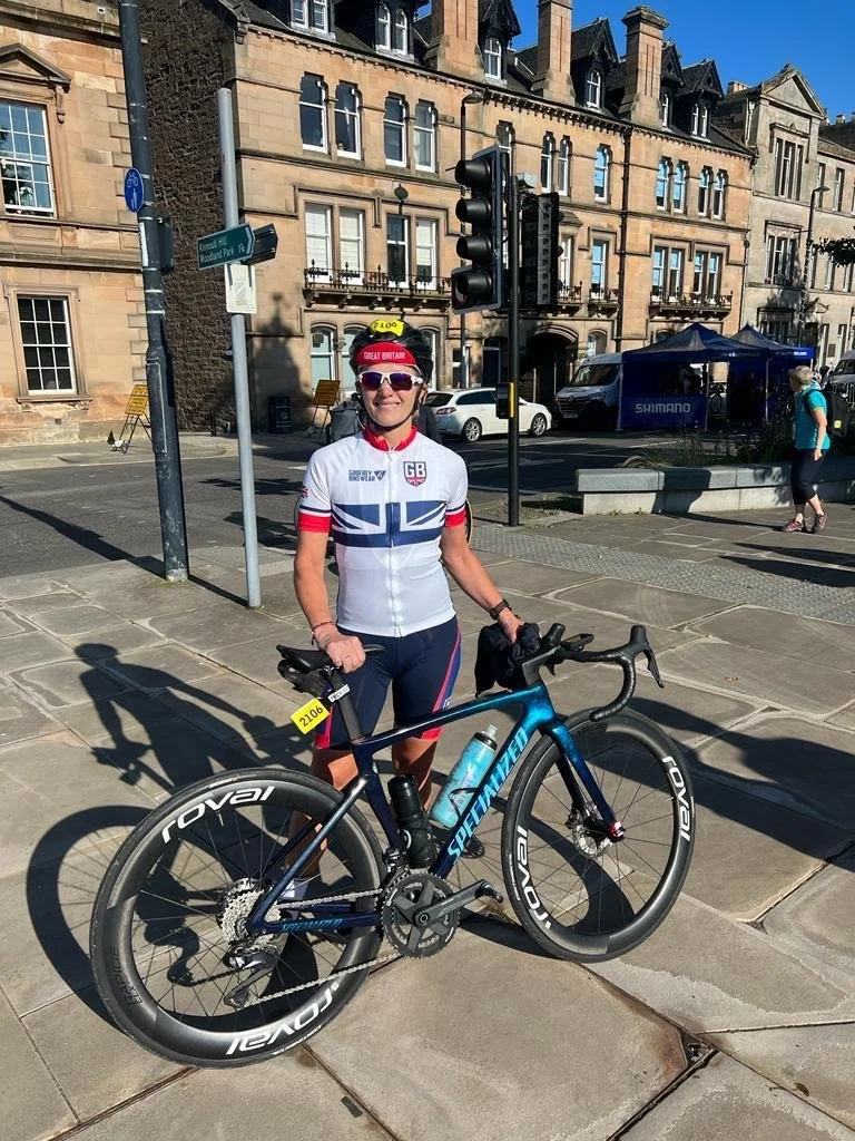 A woman standing outdoors on a city sidewalk, holding a blue Specialized road bike. She is wearing a white and red GB cycling jersey, a red and white visor, sunglasses, and black cycling shorts. There is a traffic light and historic beige stone buildings in the background.
