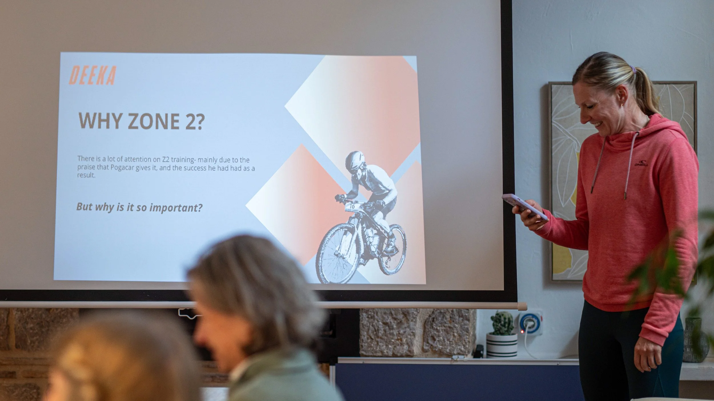 Sophie in a pink hoodie stands in front of a presentation screen, smiling as she looks at her phone. The screen displays a slide titled 'WHY ZONE 2?' with text discussing the importance of Zone 2 training and an image of a cyclist wearing a helmet.