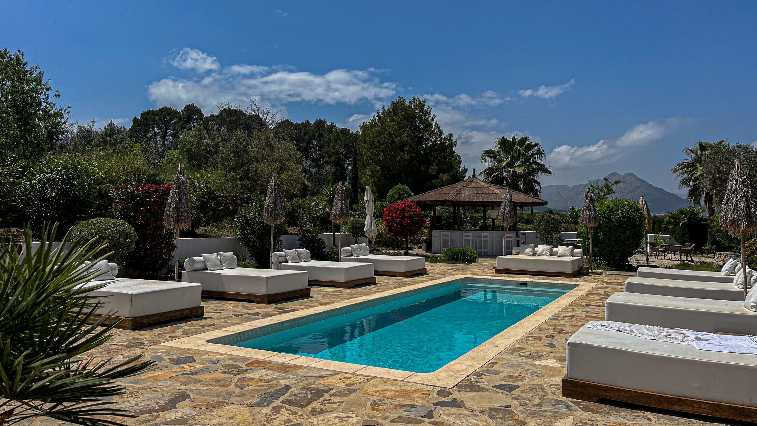 An outdoor swimming pool surrounded by white lounge chairs with pillows and straw umbrellas, lush greenery, flowers, and mountains in the background under a partly cloudy sky.