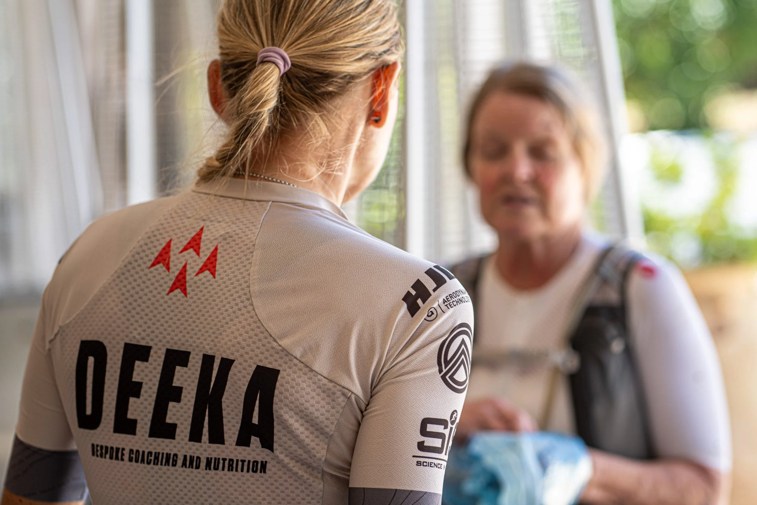 Two women in sportswear are having a conversation One woman has her back to the camera and is wearing a cycling jersey 'DEEKA' and 'BESPOKE COACHING AND NUTRITION.' The other woman faces her and is holding a blue item, wearing a backpack.