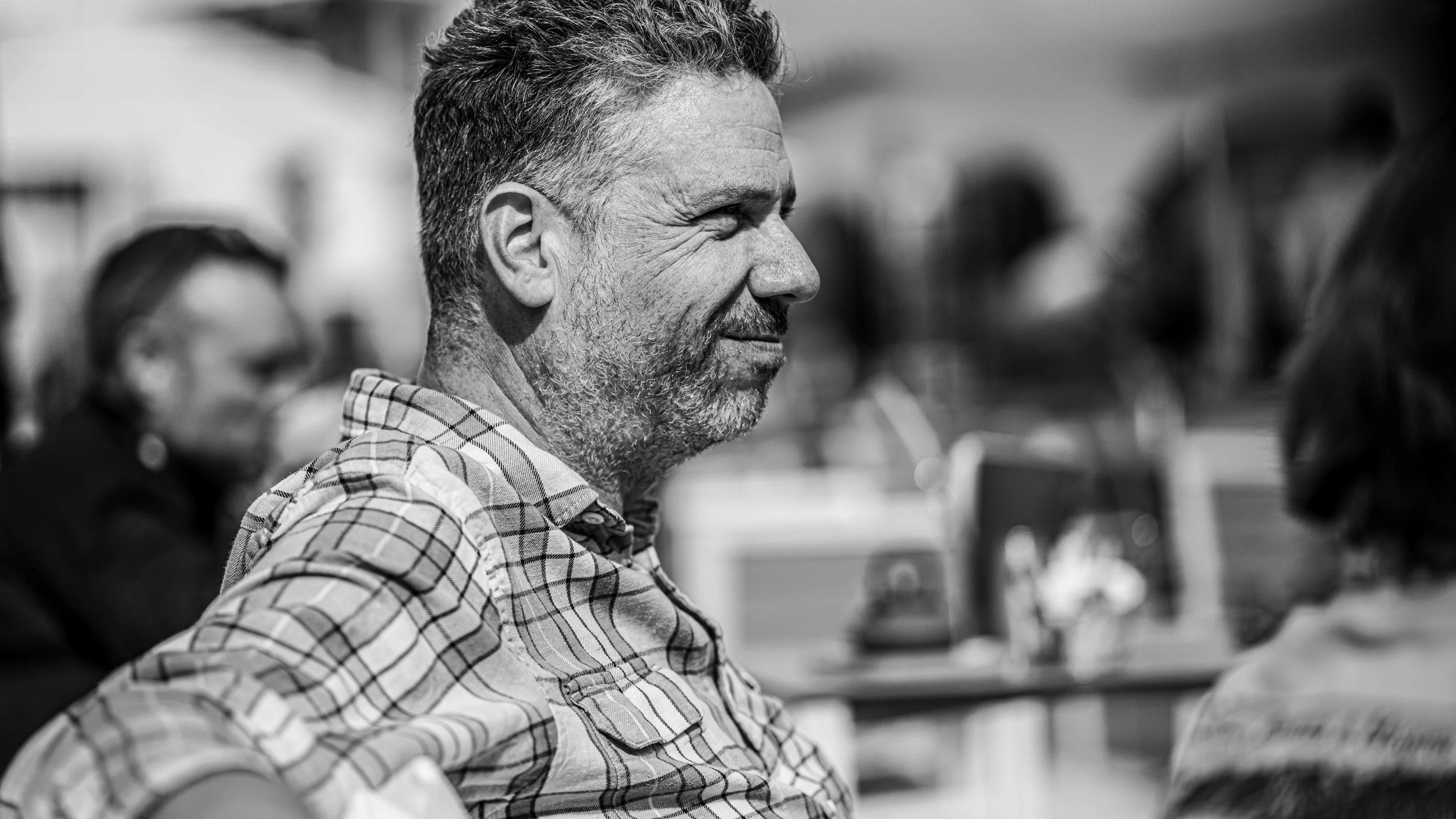 Black and white photo of a middle-aged man with short, graying hair and a beard, wearing a plaid shirt, smiling and engaging in a conversation at an outdoor event.