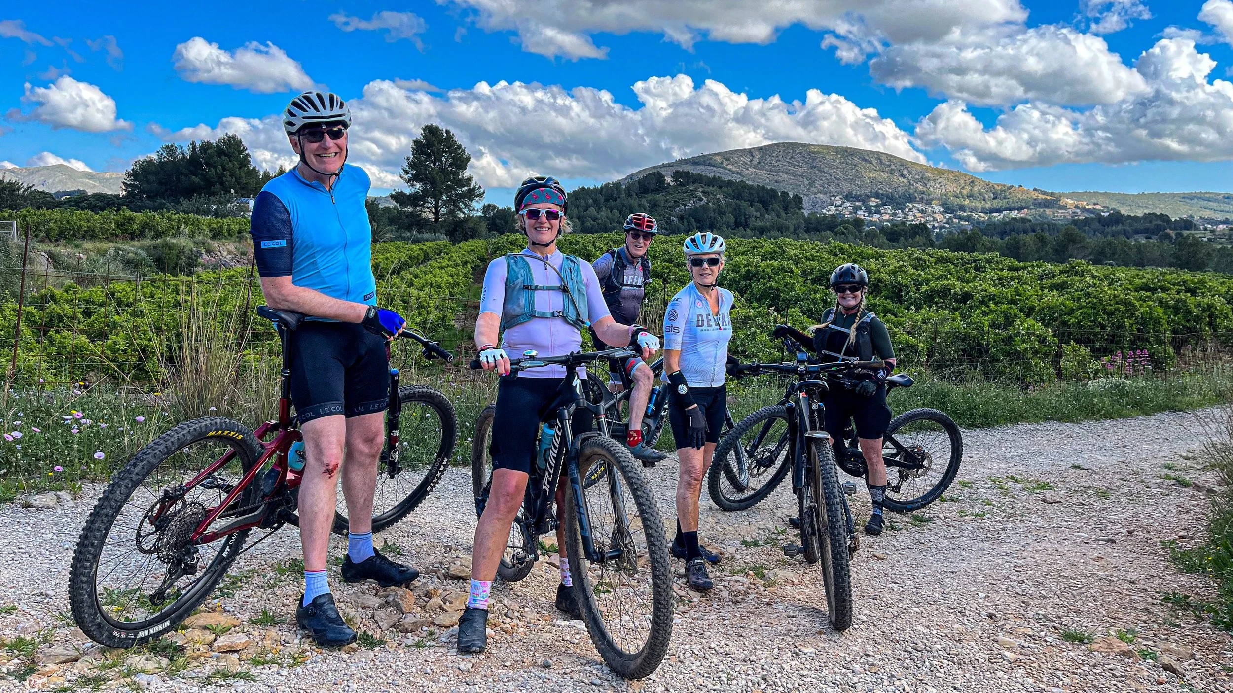 Group of five cyclists in helmets and biking gear standing with their mountain bikes on a gravel trail, with green shrubs, trees, and a mountain landscape in the background under a partly cloudy sky.