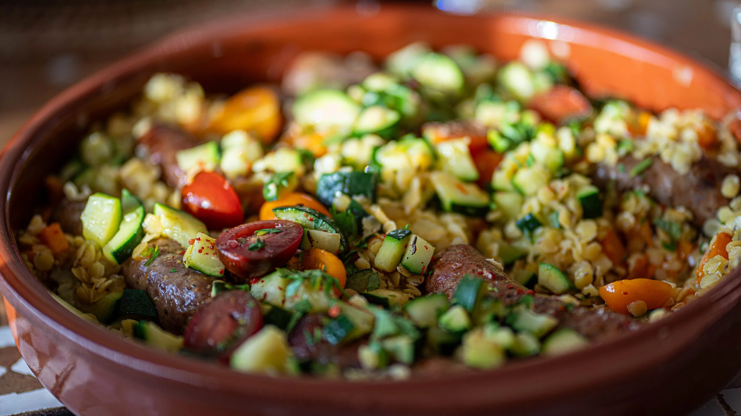 A close-up of a colorful vegetable and sausage stew in a brown ceramic bowl, containing zucchini, cherry tomatoes, carrots, and sausages.