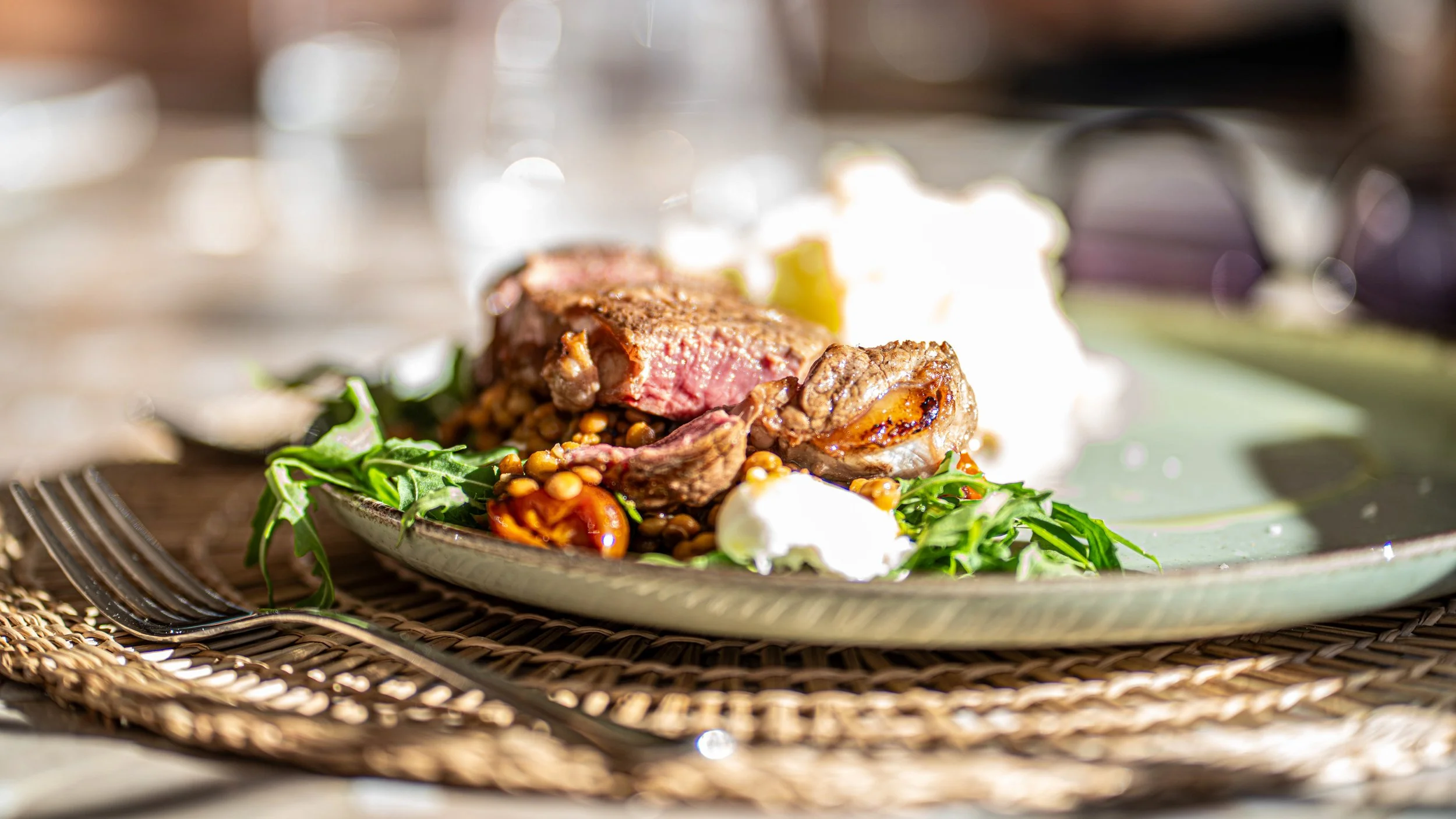 A plate of sliced steak served with greens, cherry tomatoes, and dollops of white sauce on a woven placemat, with blurred background and silver utensils nearby.