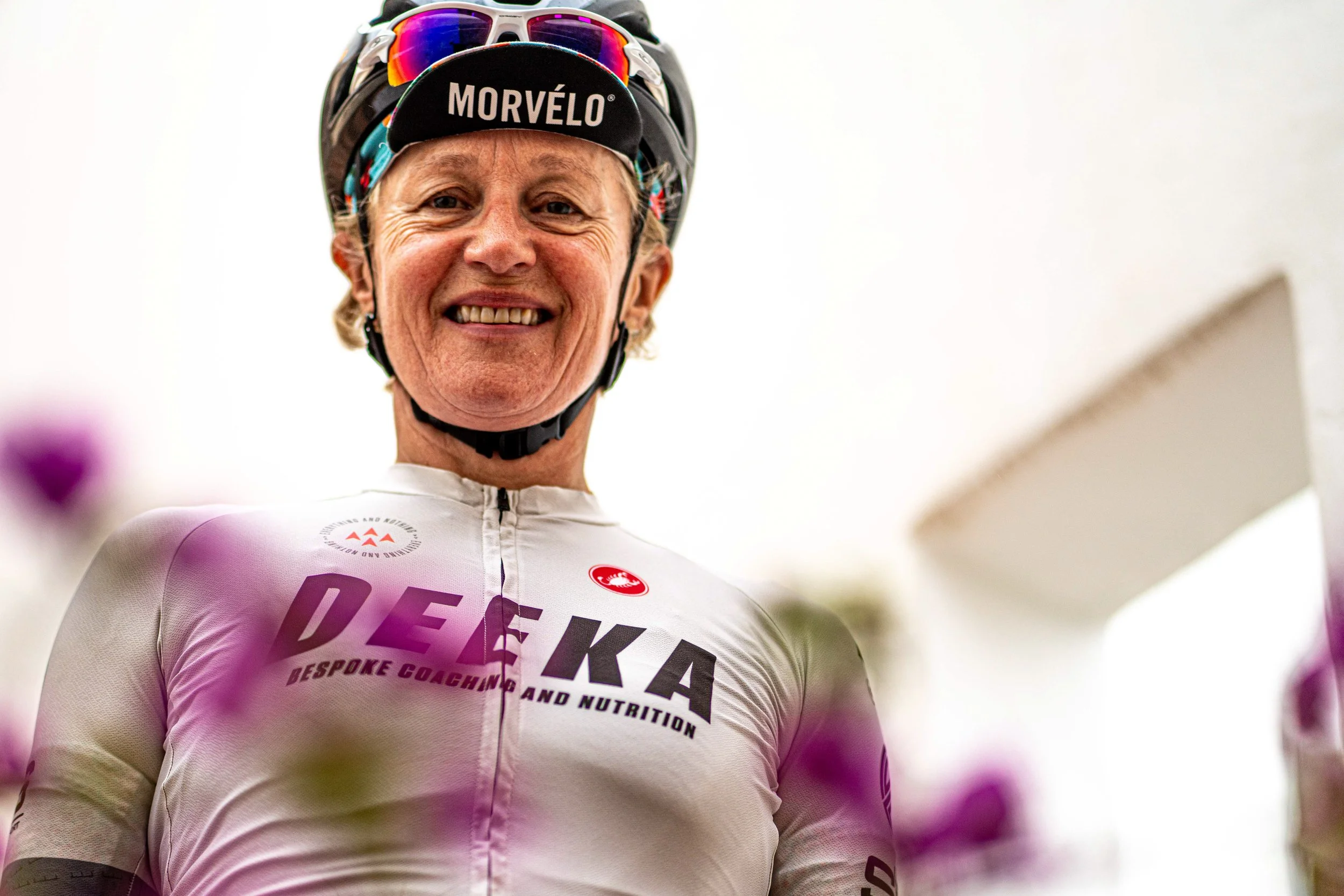 A smiling woman in a white cycling jersey with branding, wearing a black Morvelo helmet and sunglasses, surrounded by purple flowers.