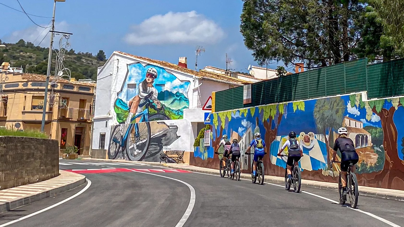 Five cyclists riding along a curved city street decorated with colorful murals on the wall to the right, featuring a girl riding a bike and an outdoor scene with trees and a building.