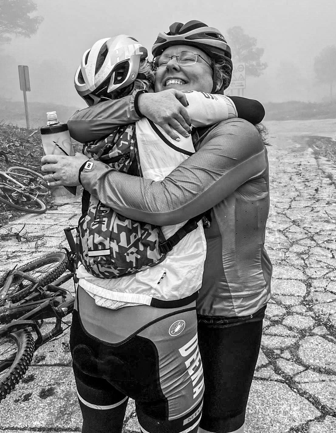 Two women in cycling gear and helmets hugging each other after a race on a cracked road in foggy weather.