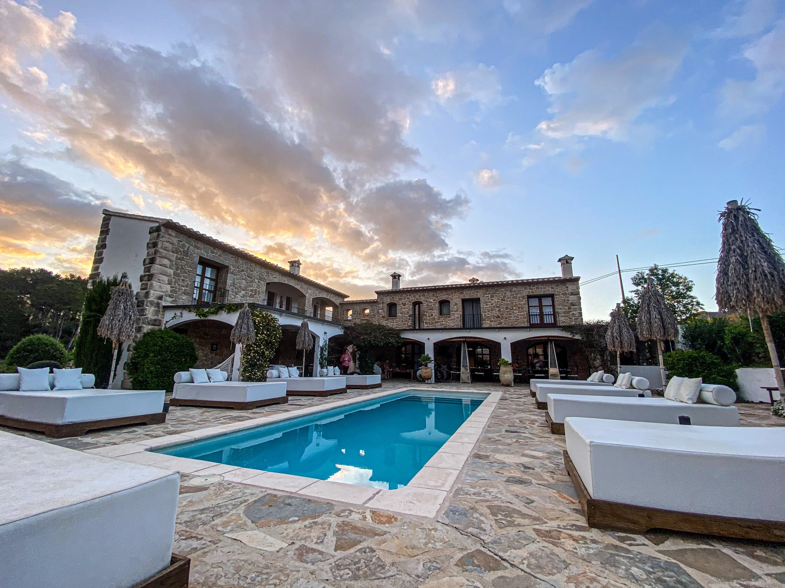 Luxury villa with stone walls and multiple levels, outdoor pool in the foreground surrounded by white lounge seating, thatched umbrellas, green bushes, and a sunset sky with clouds in the background.