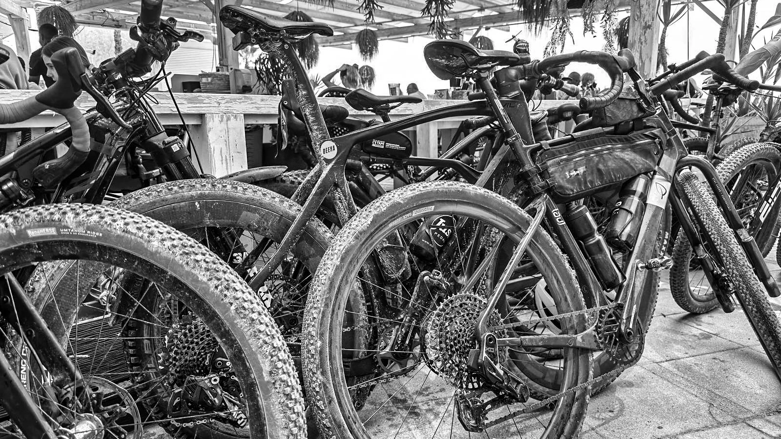 A black-and-white photo of multiple mountain bikes parked closely together indoors, with some bikes featuring thick, textured tires and others with thin, smooth tires. In the background, there are people and a wooden table.