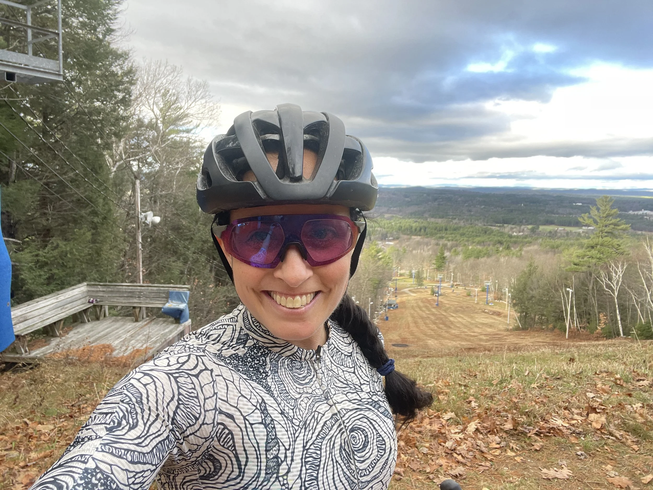 Woman in cycling gear, wearing a black helmet and pink-tinted sunglasses, smiling at the camera with a mountain bike terrain and ski lift visible in the background.