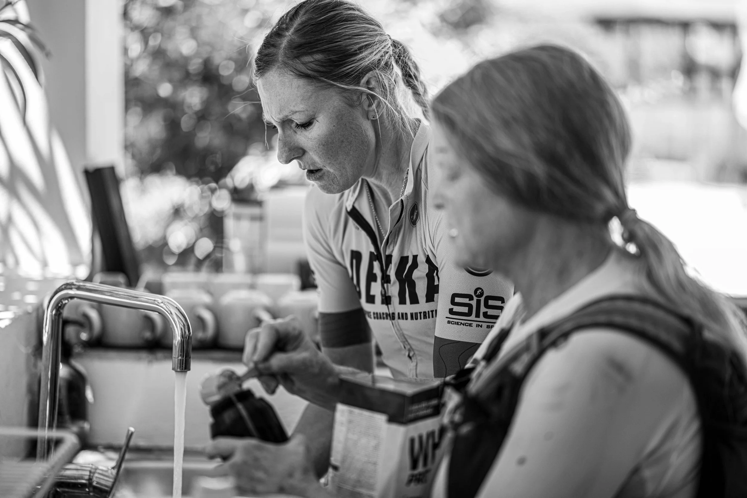 Two women at a sink, one filling a water bottle and the other observing, outdoors with blurred trees in the background, black and white photo.