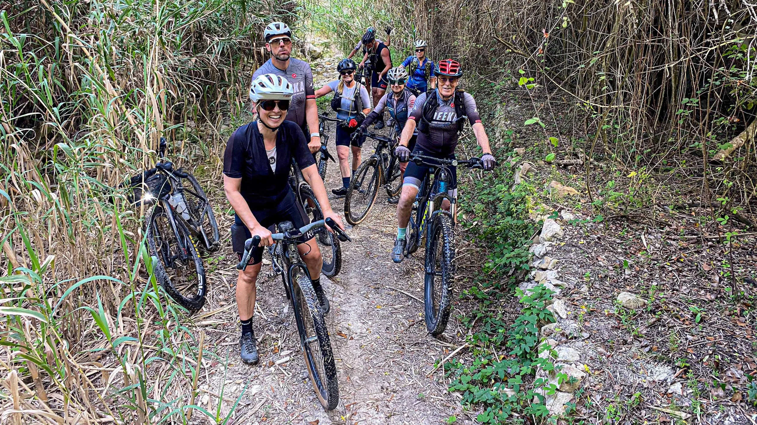 A group of nine mountain bikers, wearing helmets and sunglasses, on a dirt trail surrounded by tall grass and bushes.
