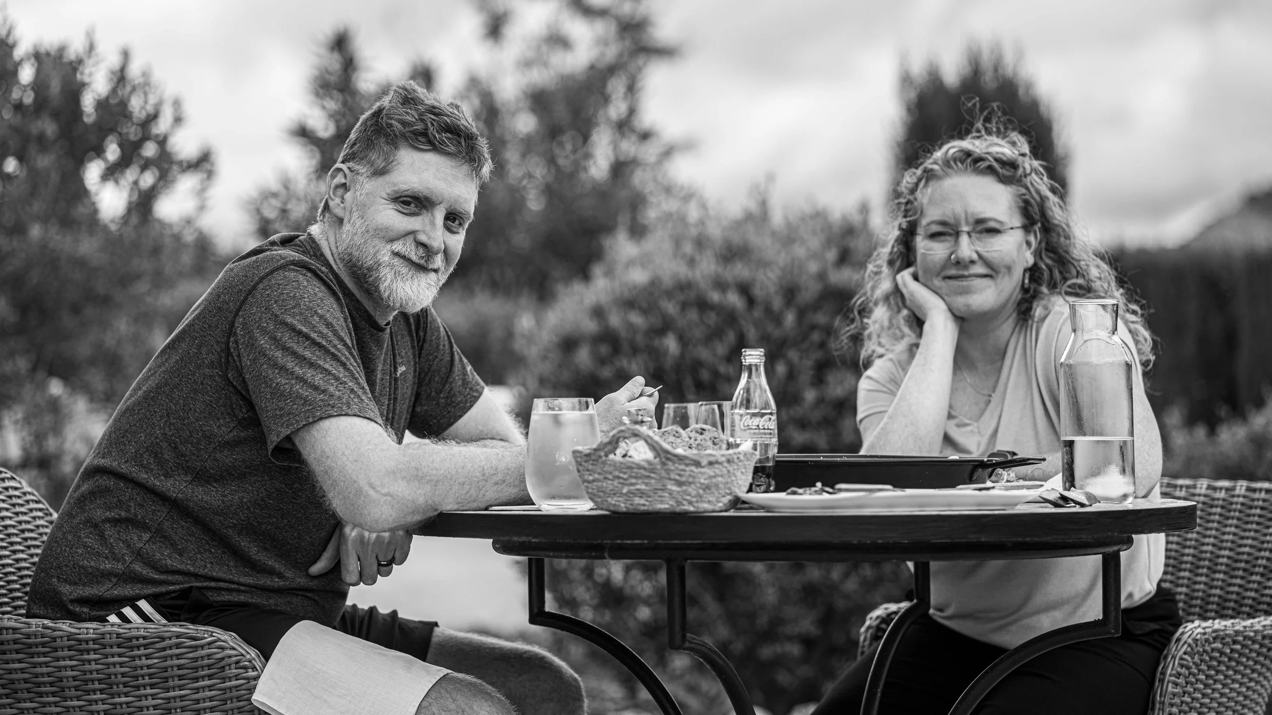 A black and white photo of a man with a beard and a woman with curly hair sitting at an outdoor table, enjoying a meal during the daytime. On the table are a basket of bread, drinks, and a pitcher of water.