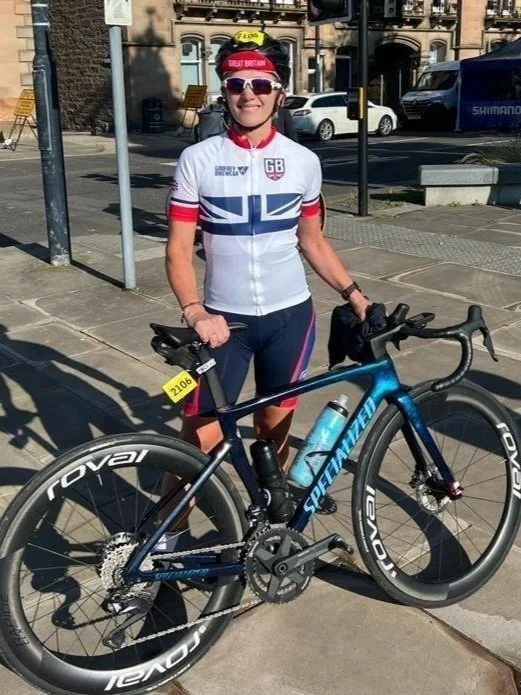 Caroline Willis cyclist,  representing team GB cycling team jersey standing on sidewalk with a blue Specialized road bike and a water bottle, wearing sunglasses, a red headband, and a yellow identification tag, in an urban setting.