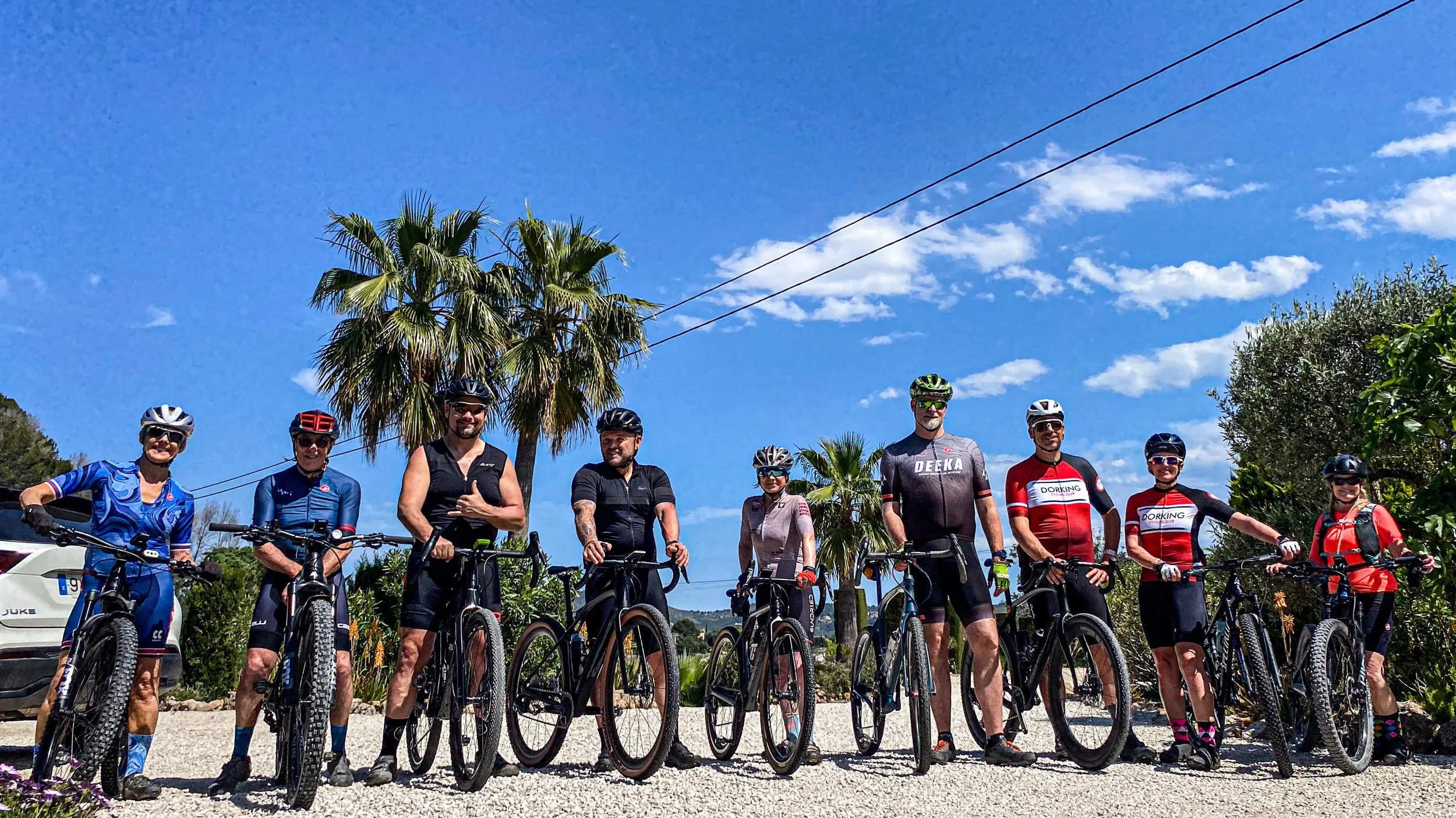 Group of nine cyclists standing with their bikes outdoors on a sunny day, with palm trees and bushes in the background, and blue sky with some clouds.