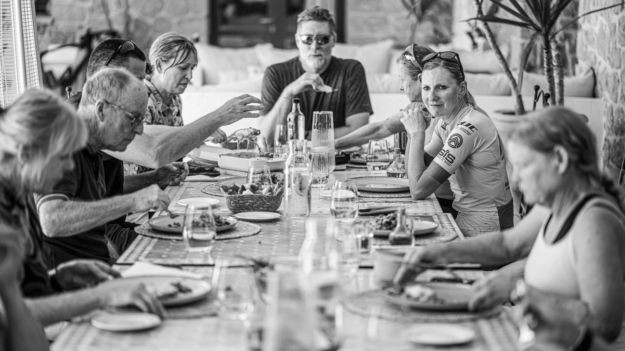 A black and white photo of a group of people sitting at a long dining table, eating and conversing at DEEKA Elevate cycling camp 2025. The table is set with glasses, plates, and a basket of food.