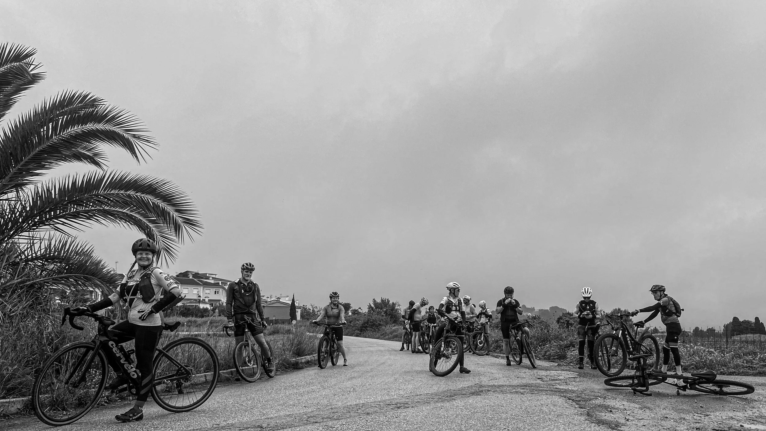 Group of cyclists on a rural road, some standing with bicycles, one lying on the ground, under cloudy sky, with buildings and trees in the background.